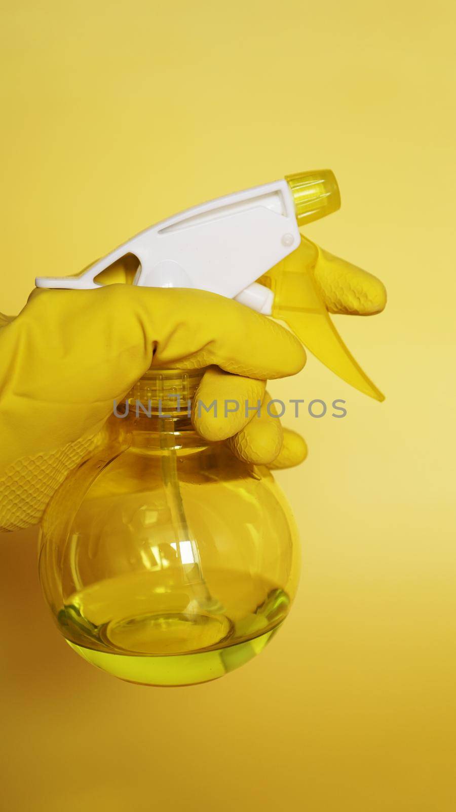 Hand in yellow rubber glove holding plastic spray bottle with cleaning detergent on yellow background - vertical