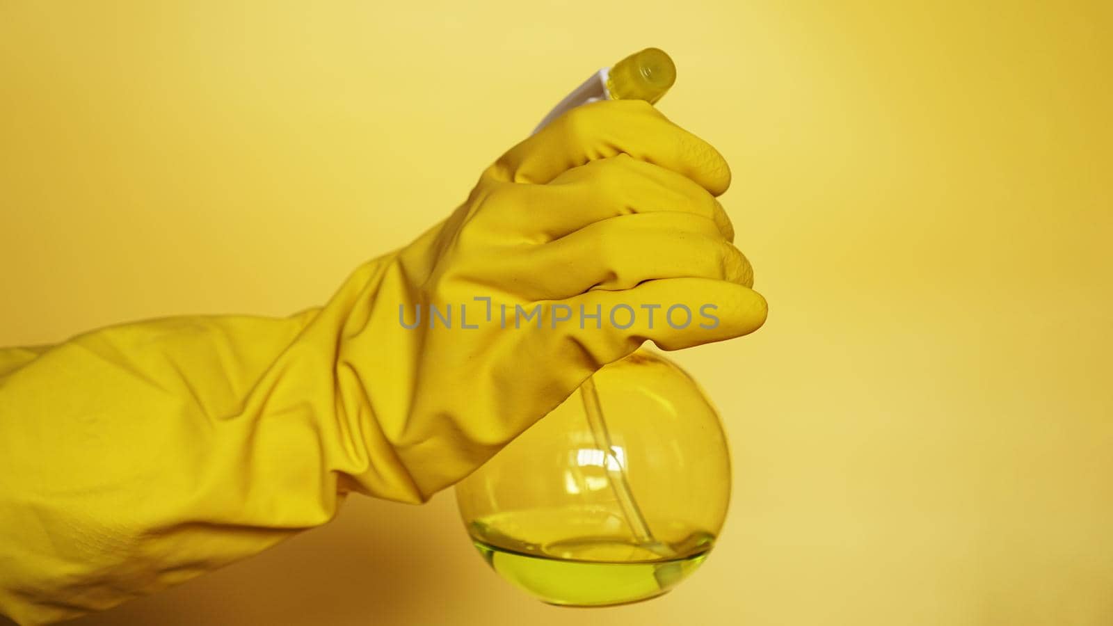 Hand in yellow rubber glove holding plastic spray bottle with cleaning detergent on yellow background