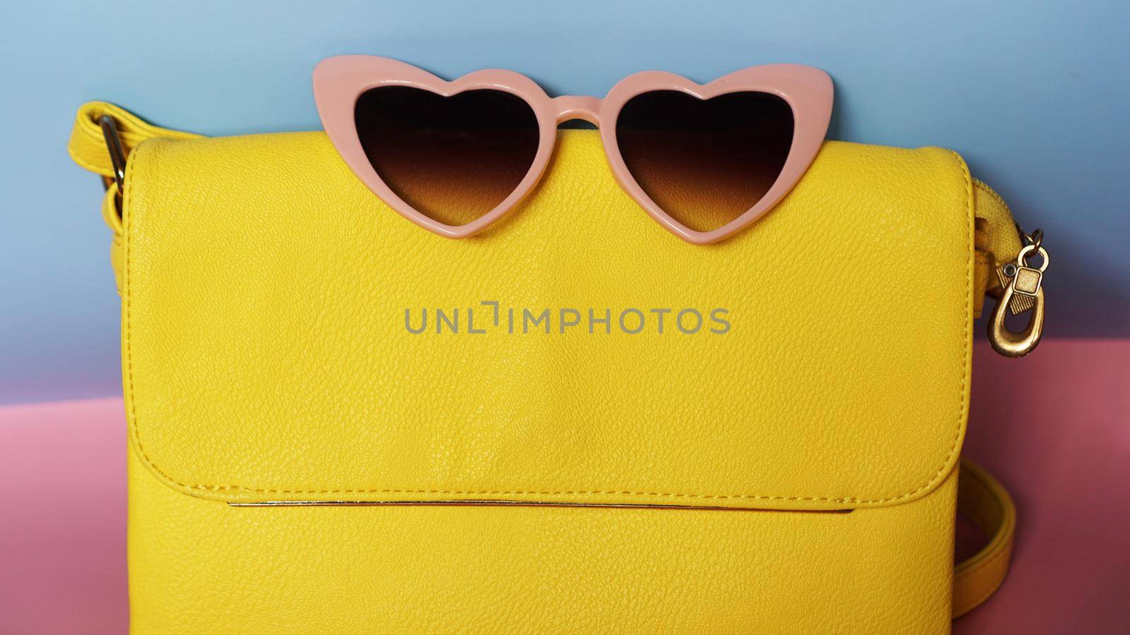 Yellow bag and heart-shaped sunglasses on pink and blue background by natali_brill