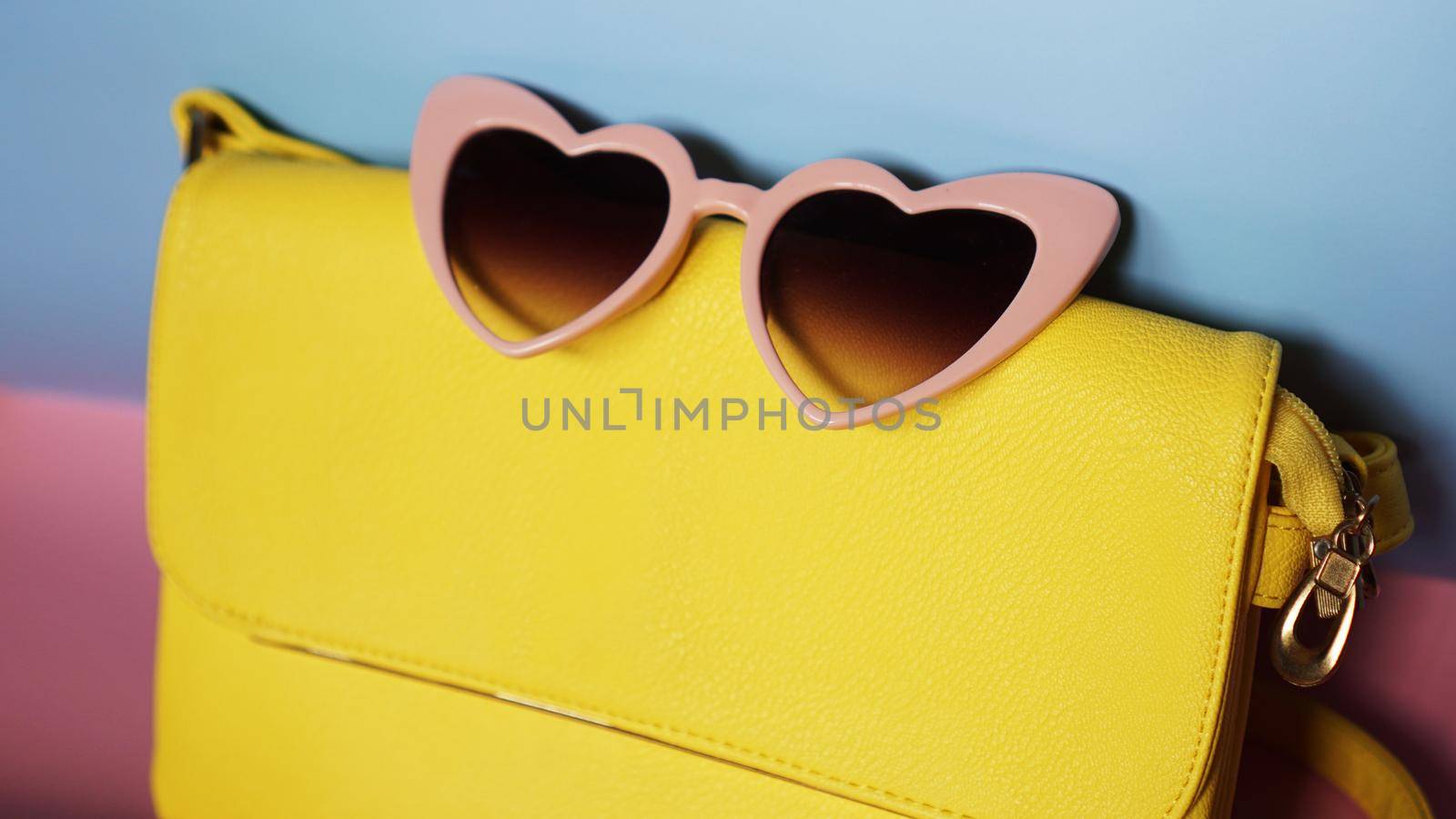 Yellow bag and heart-shaped sunglasses on pink and blue background by natali_brill