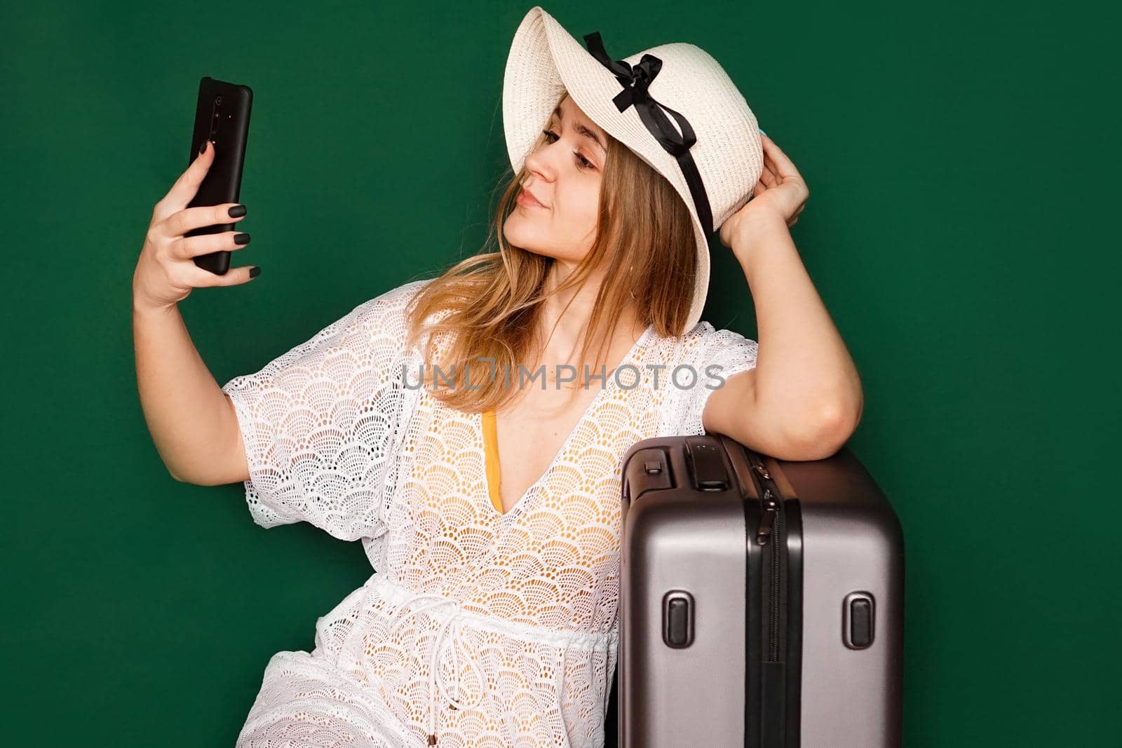 Elegant european woman taking selfie before vacation. Indoor shot of a lovely girl in a white dress sitting near her suitcase, green background