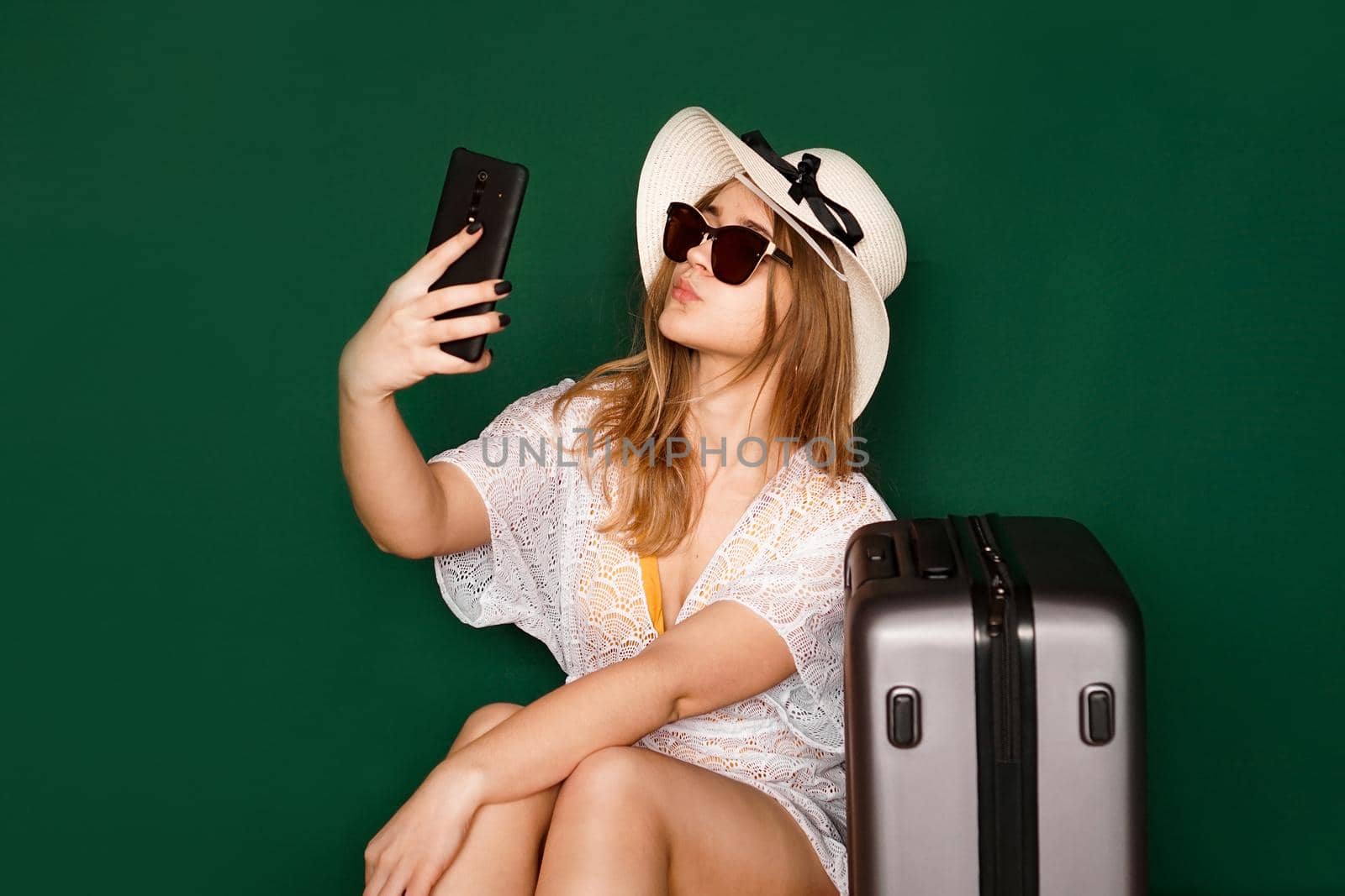 Elegant european woman taking selfie before vacation. Indoor shot of a lovely girl in a white dress sitting near her suitcase, green background