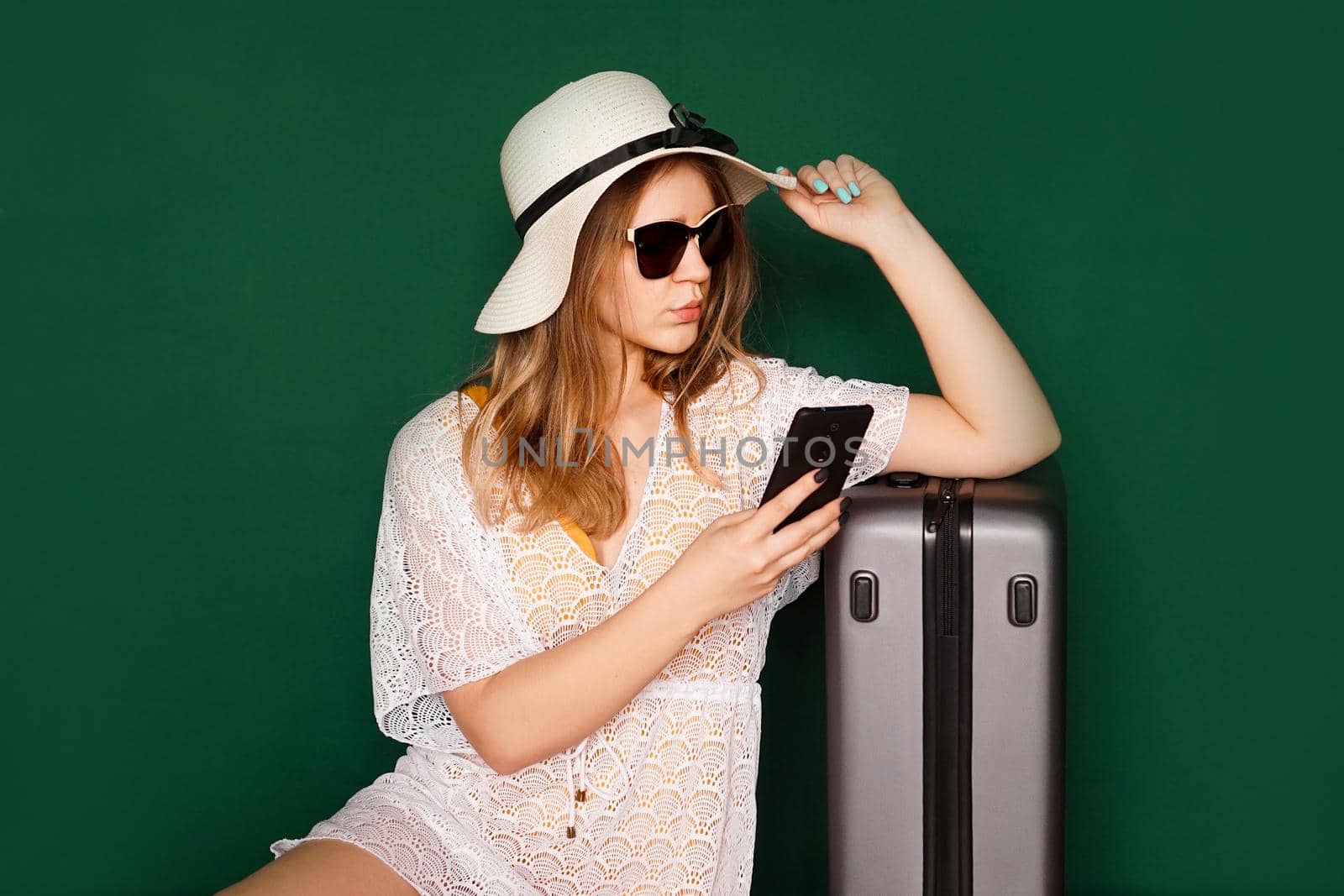 Beautiful girl in a white dress and a hat sits near a suitcase. Waiting for departure. Girl with a phone on green background