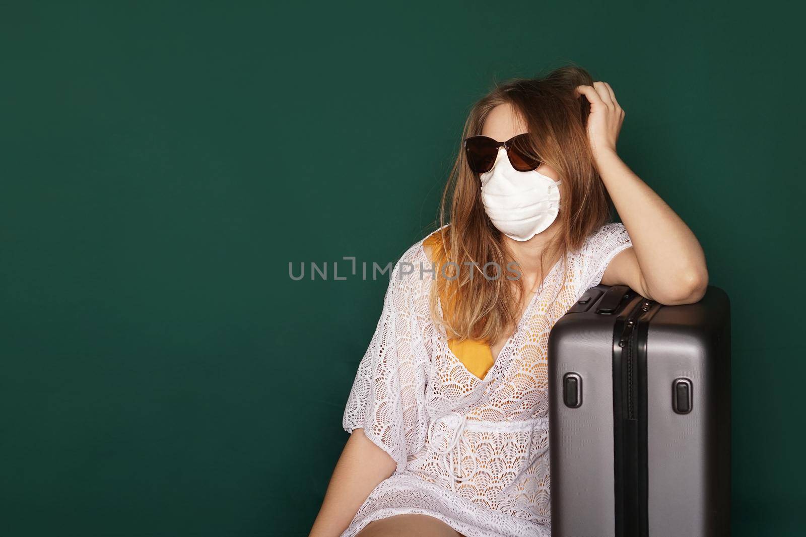 Beautiful girl in a white summer dress with a suitcase. Medical mask on the face. Closed borders during coronavirus. Tourist on a green background
