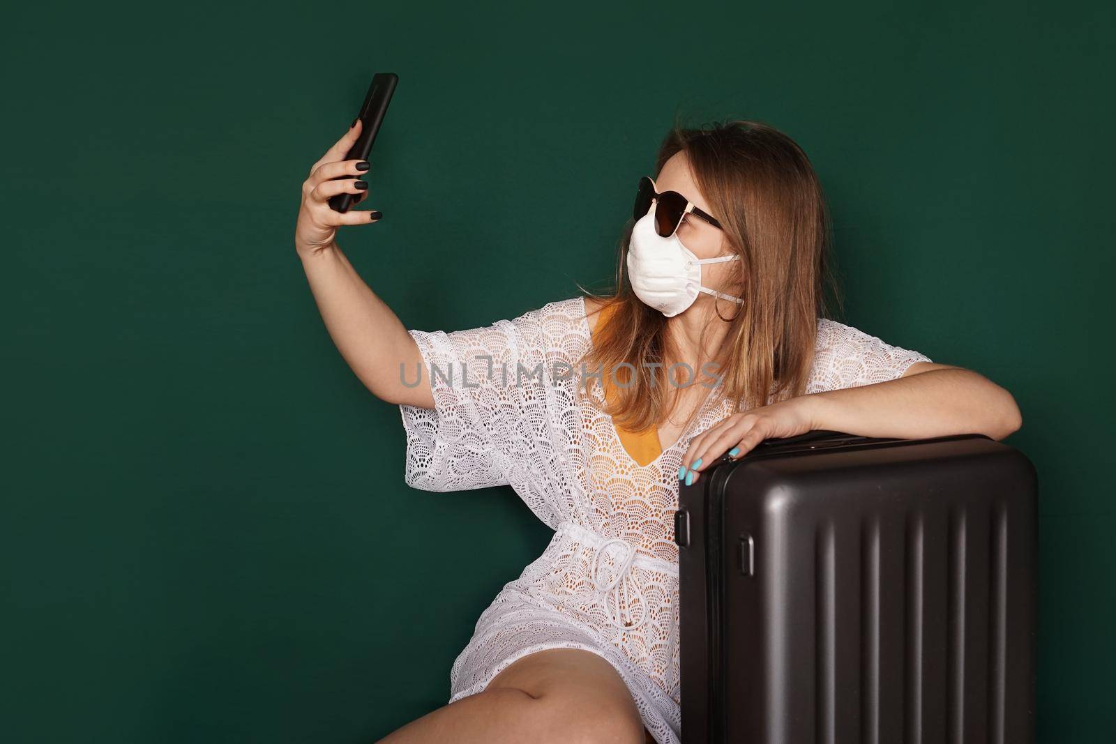 Beautiful girl in a white summer dress with a suitcase taking selfie . Medical mask on the face. Closed borders during coronavirus. Tourist