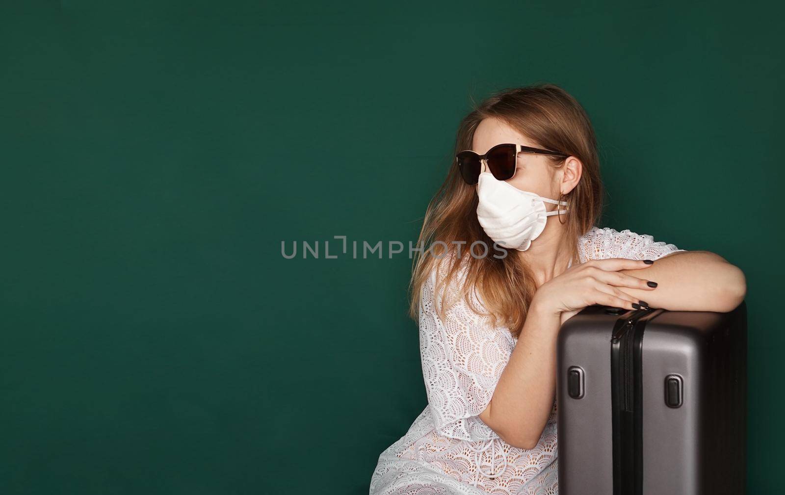 Beautiful girl in a white summer dress with a suitcase. Medical mask on the face. Closed borders during coronavirus. Tourist on a green background