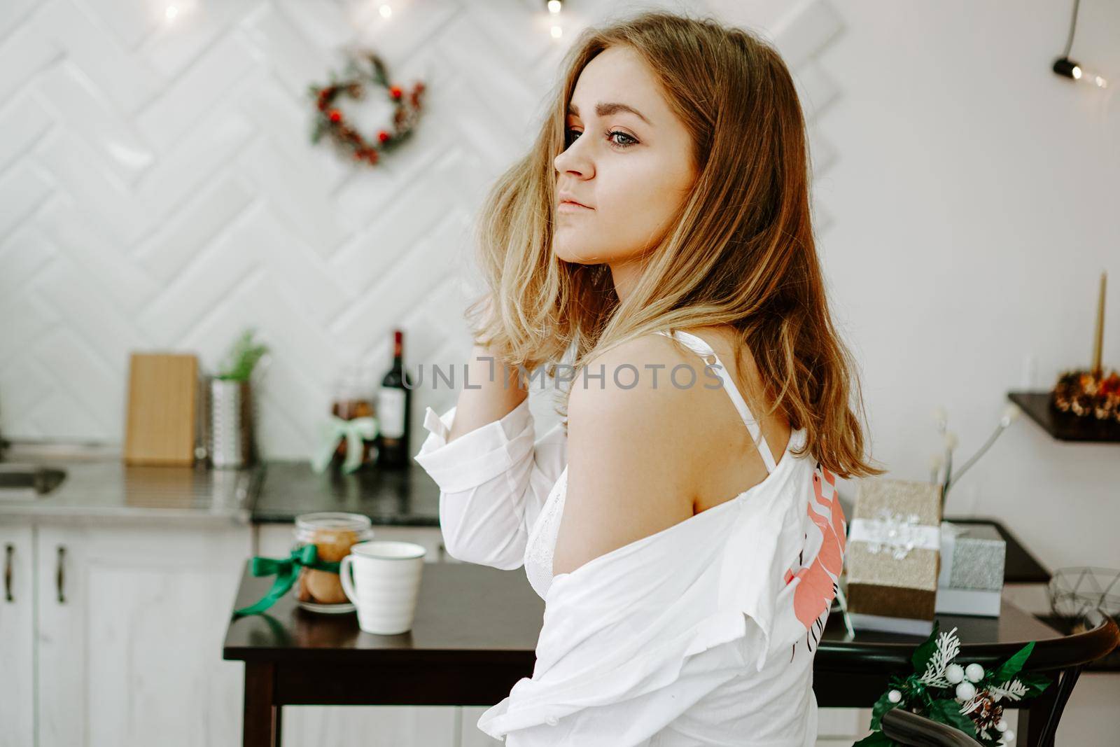 A girl in a long white shirt stands in the kitchen. The white kitchen is decorated with Christmas decorations and garlands