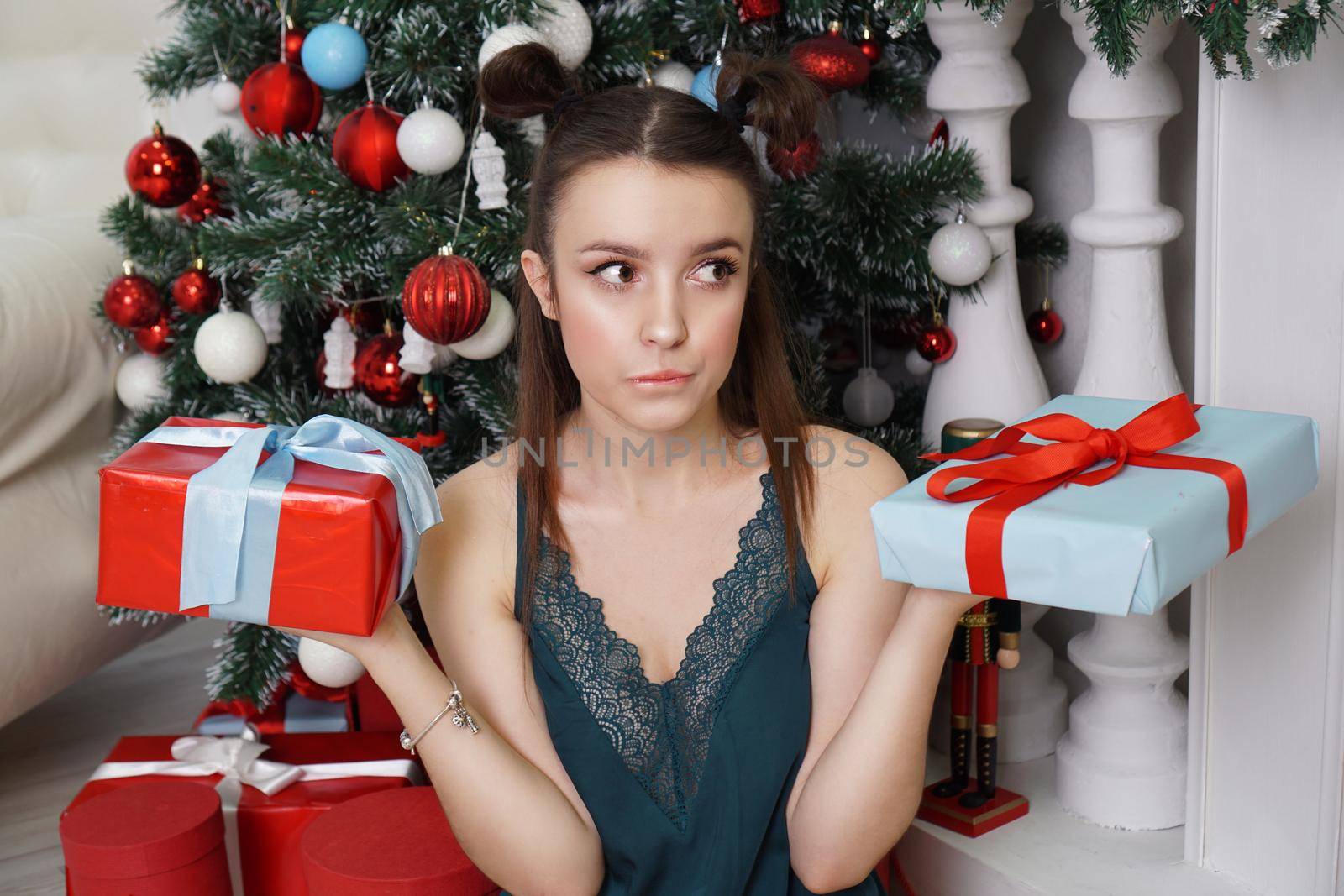 Happy girl in a green peignoir and funny hairstyle holding two gift boxes, not knowing which gift to choose to give a Christmas tree, decorated room