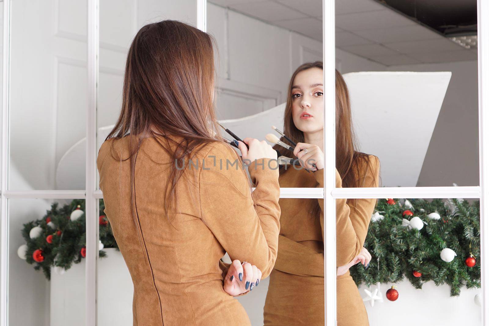 Makeup artist with long dark hair holding a powder brush, haughty expression, reflection in the mirror, christmas decor behind