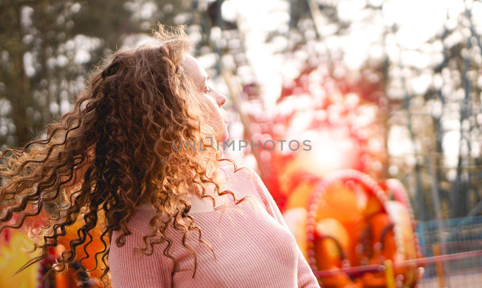 Girl chilling in amusement park in weekend morning. Laughing good-humoured female model with curly hair standing near carousel.