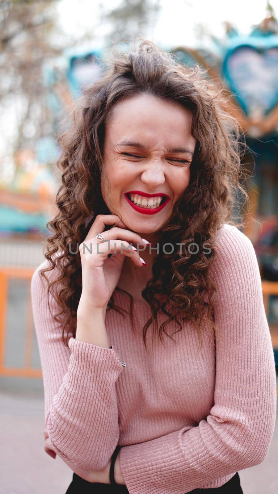 Girl chilling in amusement park in weekend morning. Laughing good-humoured female model with curly hair standing near carousel.