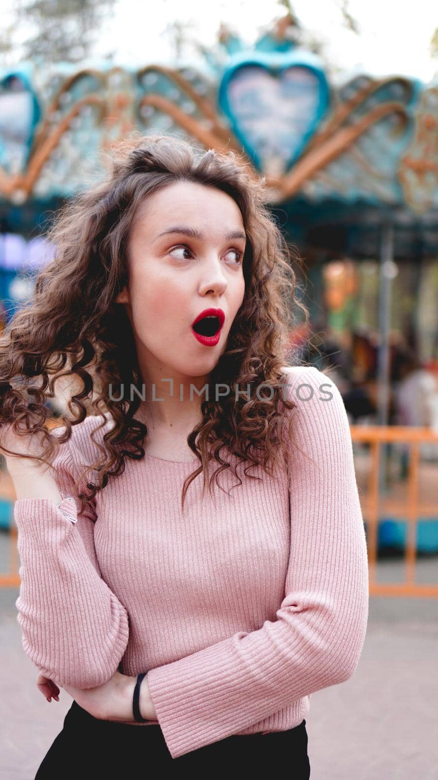 Girl chilling in amusement park in weekend morning. Laughing good-humoured female model with curly hair standing near carousel.