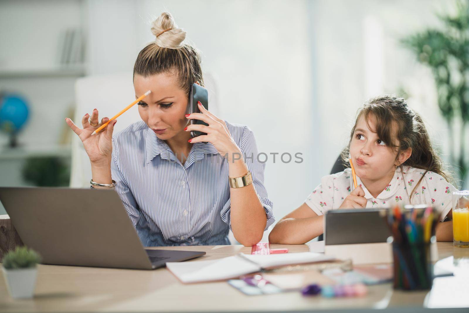 Busy single mother using laptop and talking on her smartphone while her cute daughter learning at home.