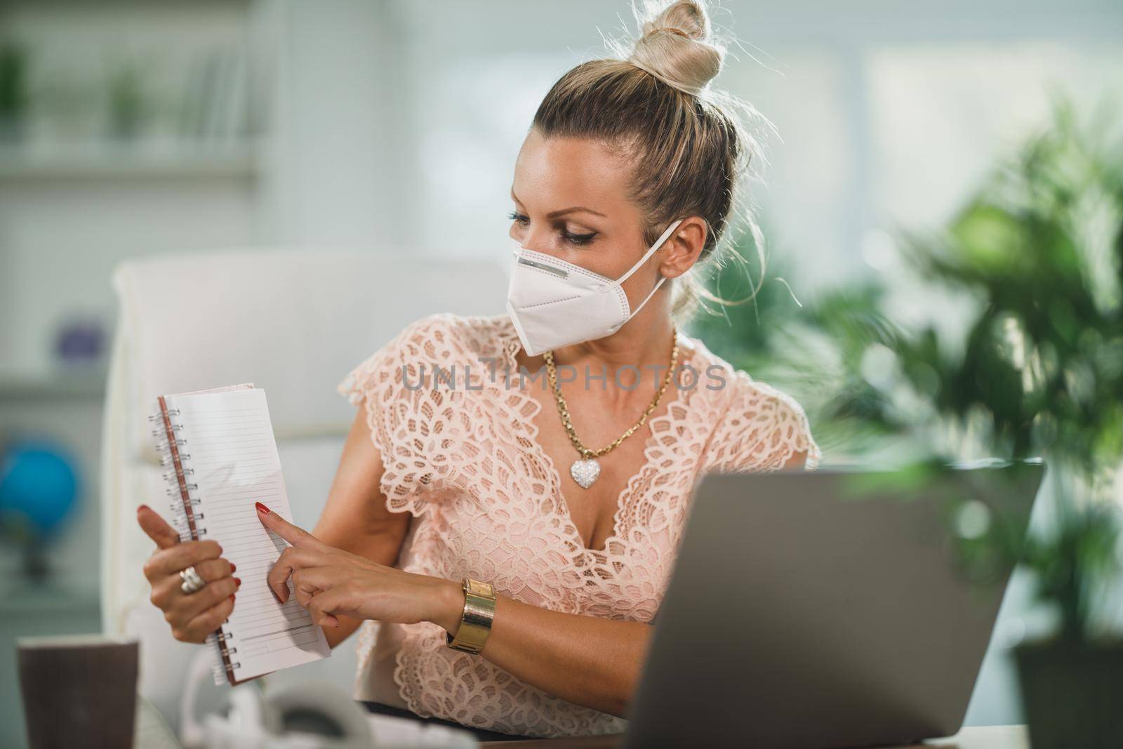 An attractive business woman with N95 protective mask sitting alone in her home office and working online at her laptop.