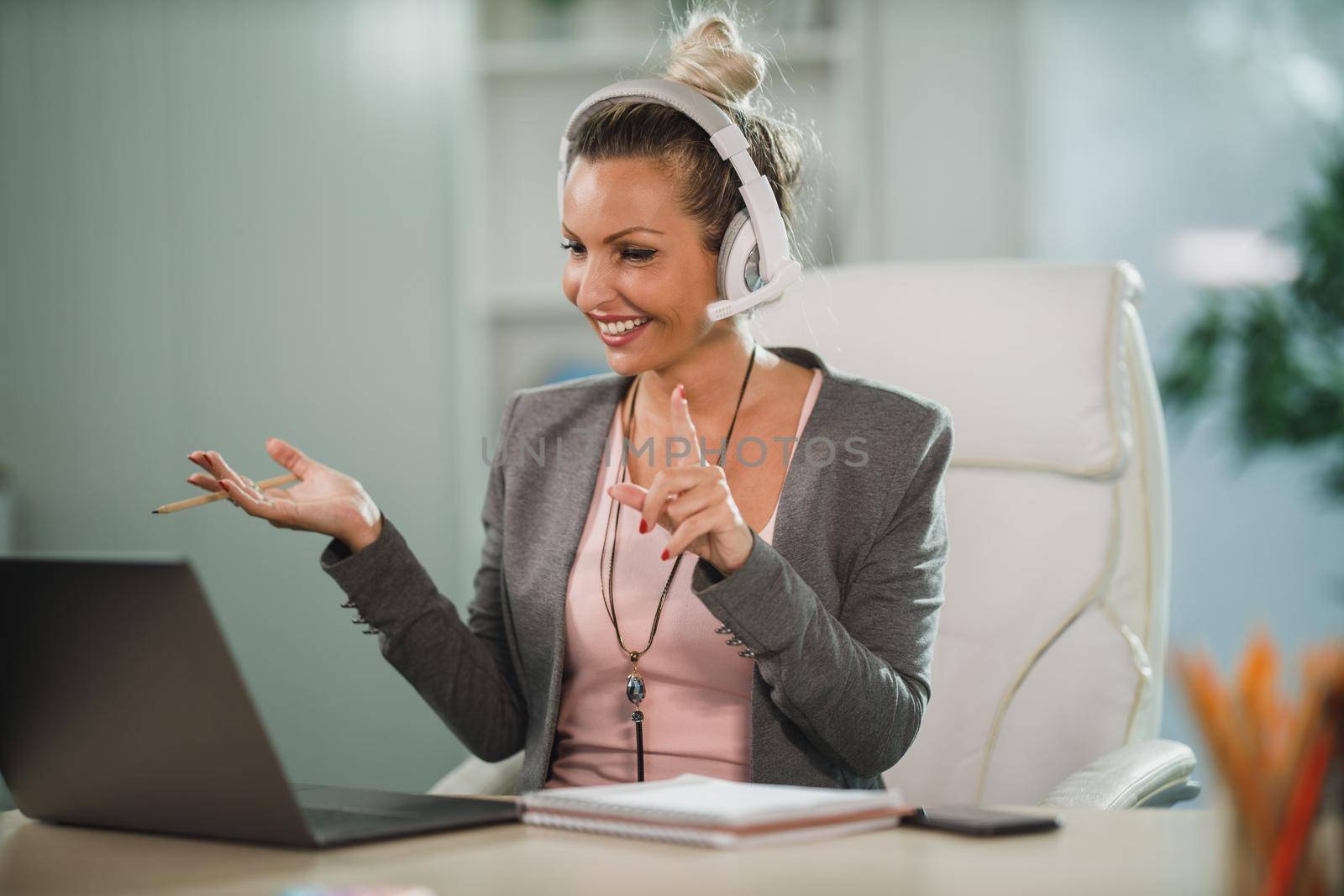 An attractive business woman with headphones making video call at her laptop and teaching students in her home office.