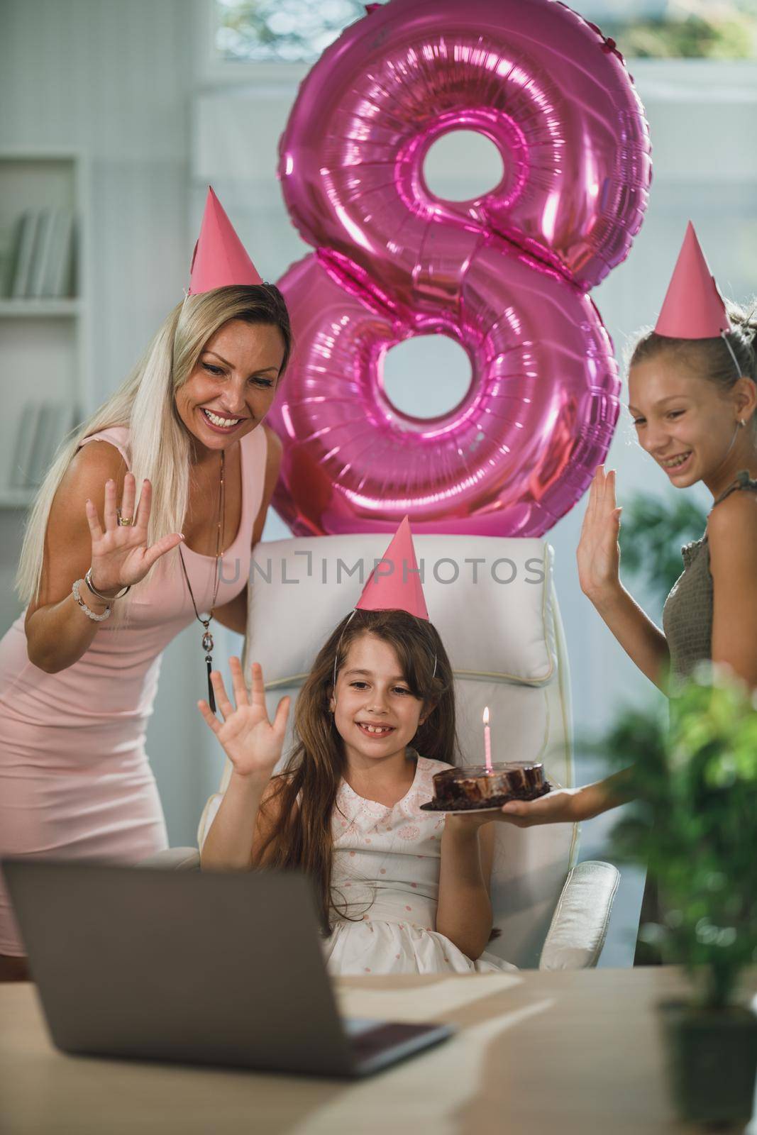 Beautiful young mom with her cute daughters have video call with friends while birthday celebration at home during pandemic isolation.