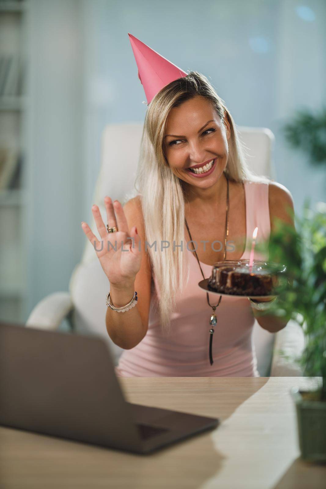 Alone attractive woman holding a birthday cake and have video call with friends while birthday celebration at home during pandemic isolation.