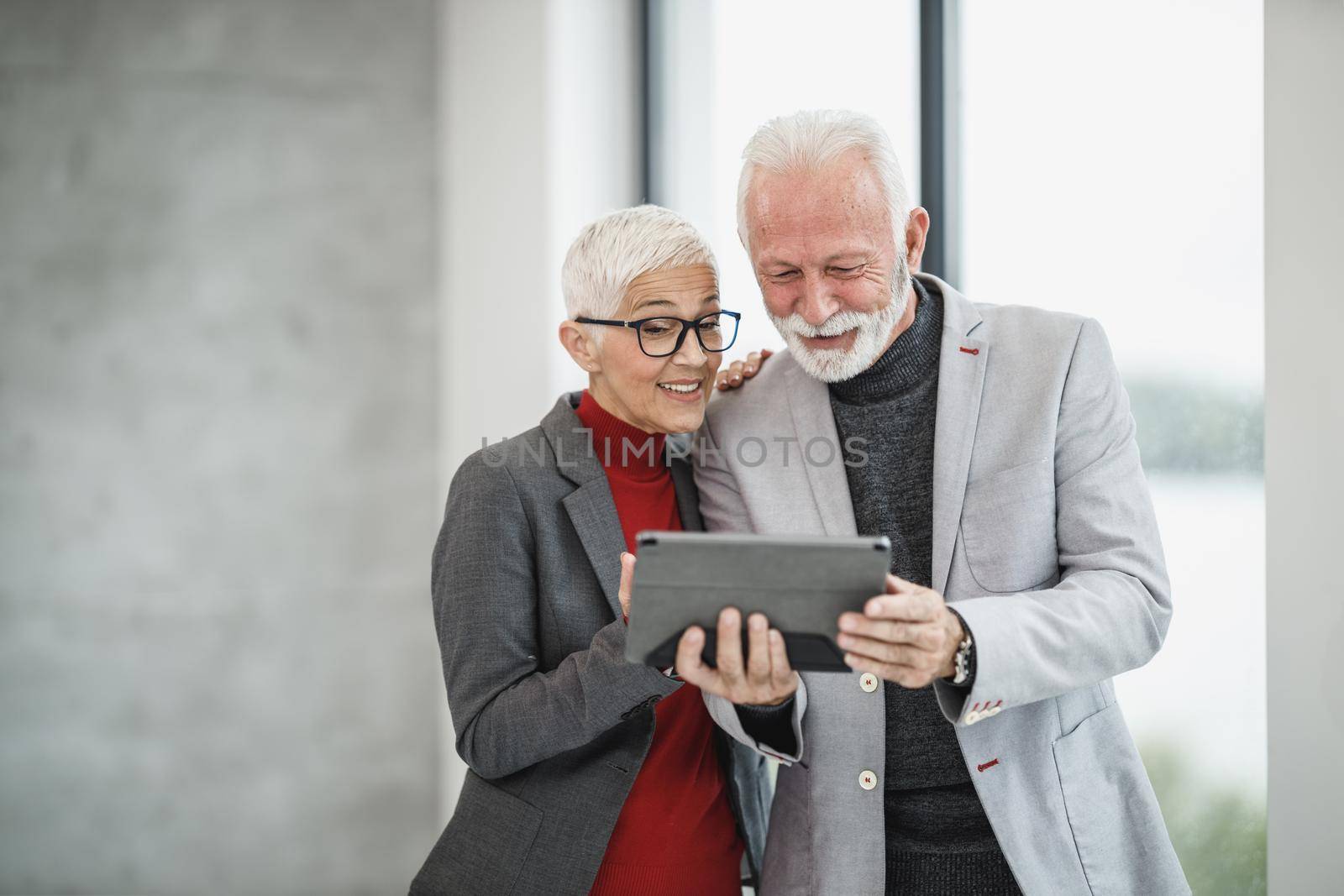 A successful senior businessman and his smiling female colleague making video call on a digital tablet in a modern office.