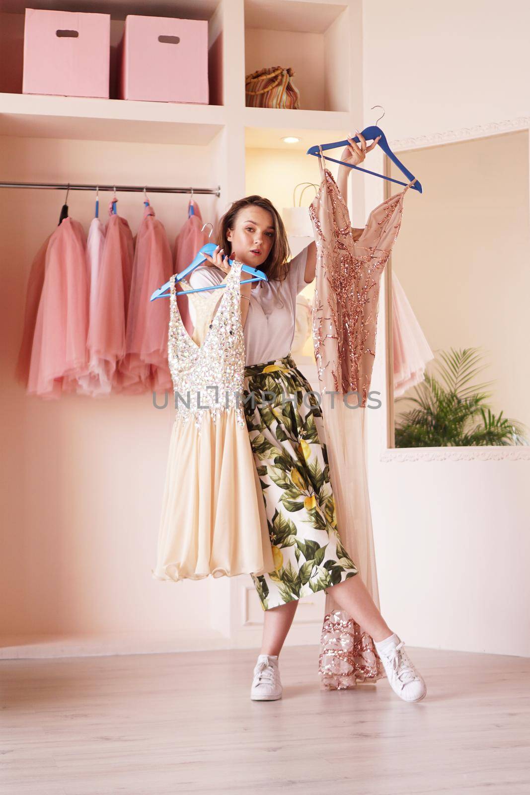 Young woman standing near wardrobe, holding dress on hangers, trying to decide what to wear. Pretty female choosing clothes in dressing room.