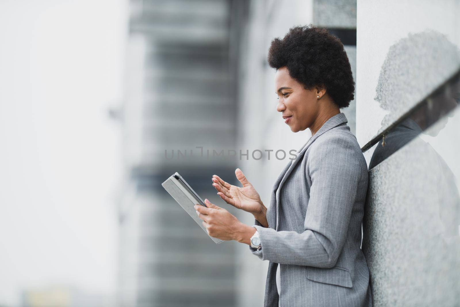 A successful black business woman making video call on a digital tablet during quick break in front a corporate building.