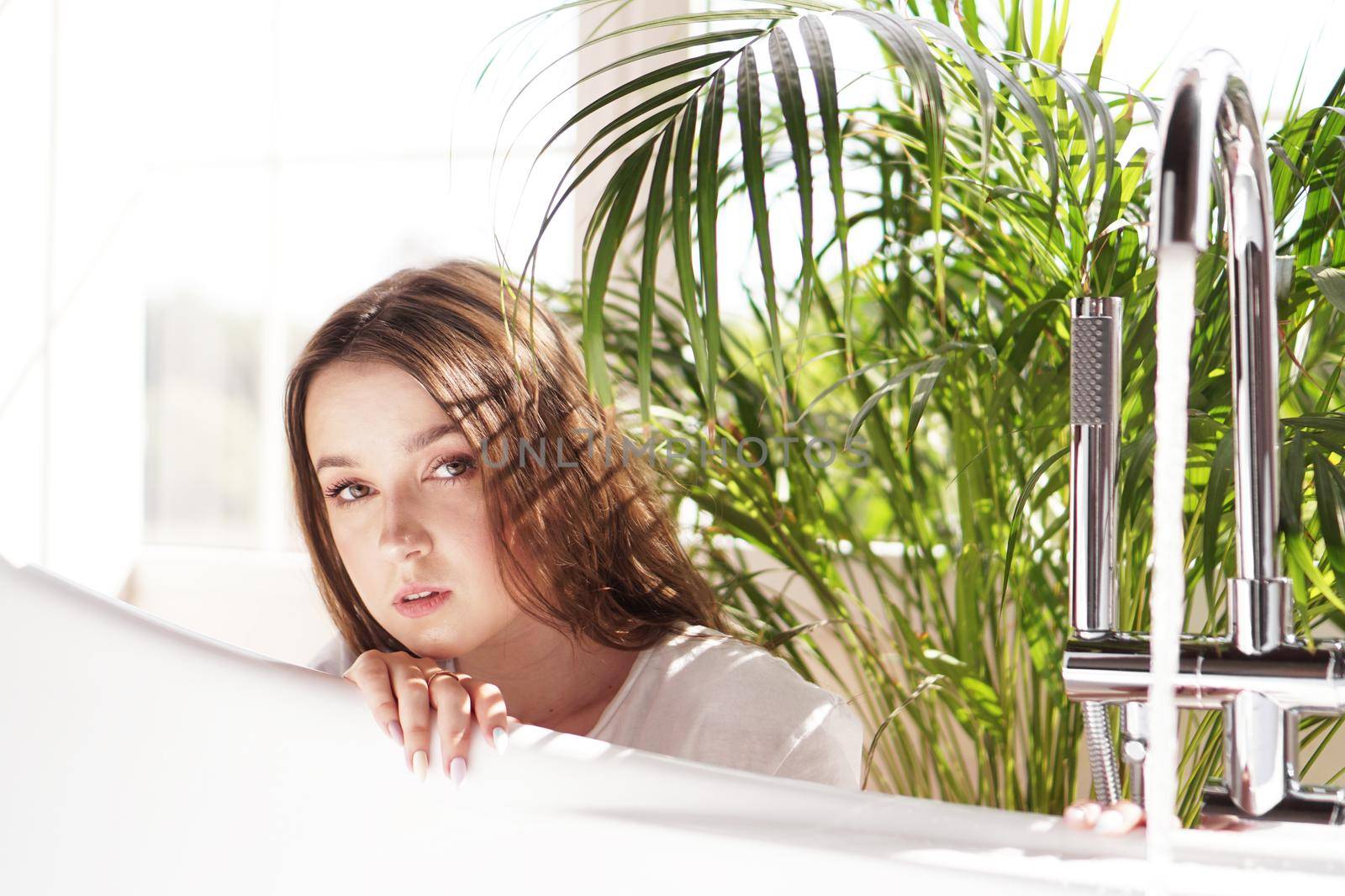 Think positive. Girl in a bright bathroom with a green plant. A happy woman waits until the bathtub is filled with water