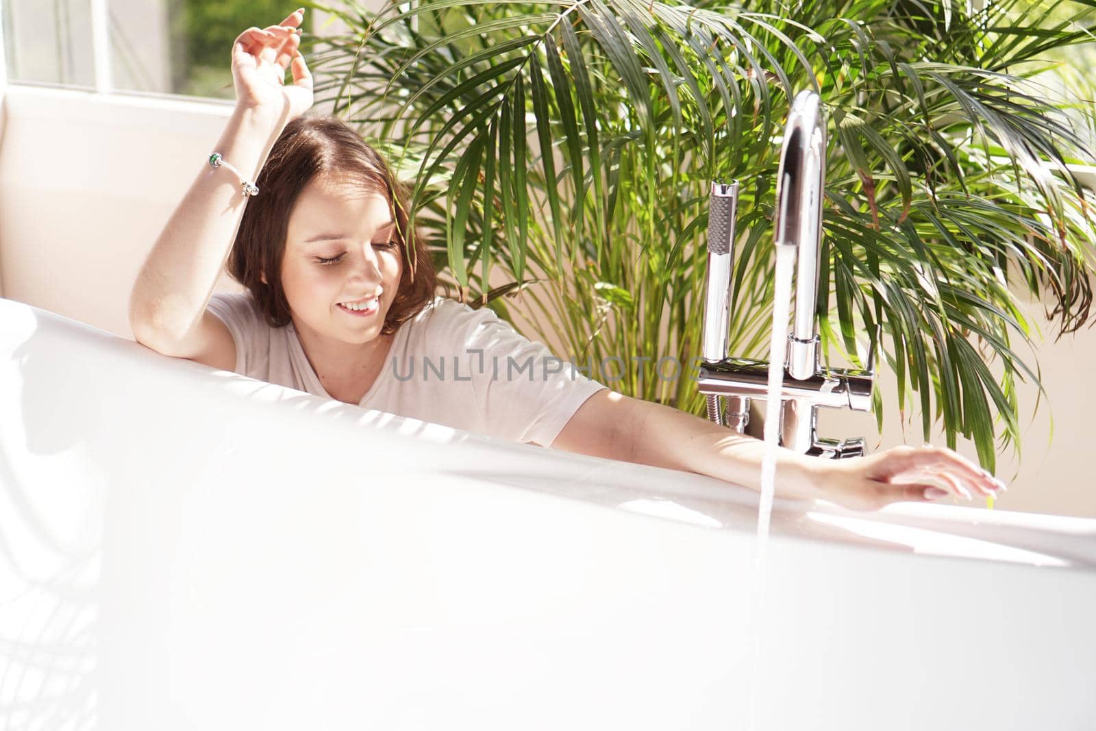 Think positive. Girl in a bright bathroom with a green plant. A happy woman waits until the bathtub is filled with water