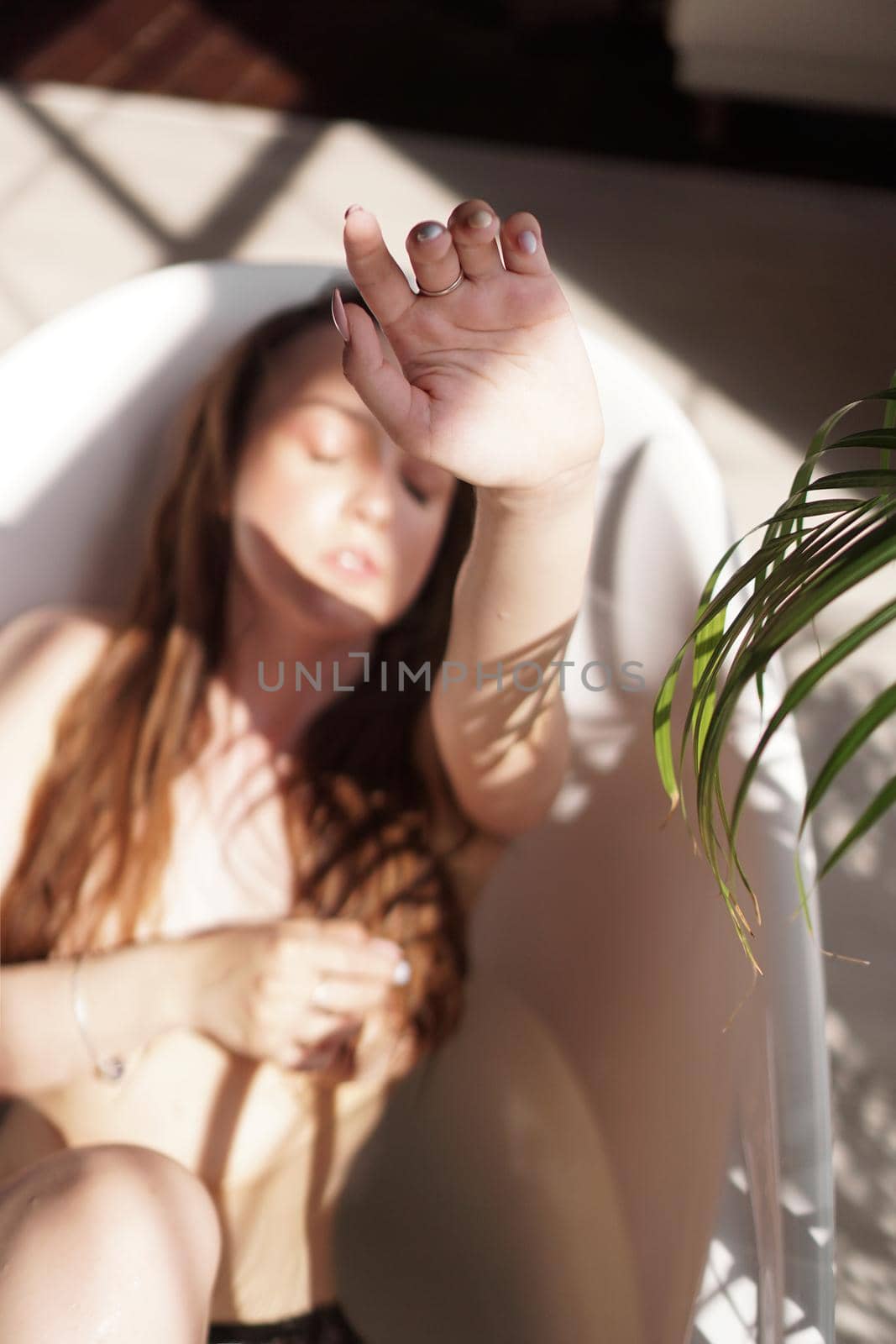 Young attractive woman relaxing in bath - plant in bathroom - sunny day. The model raises the hand - focus on the fingers. Blurred background