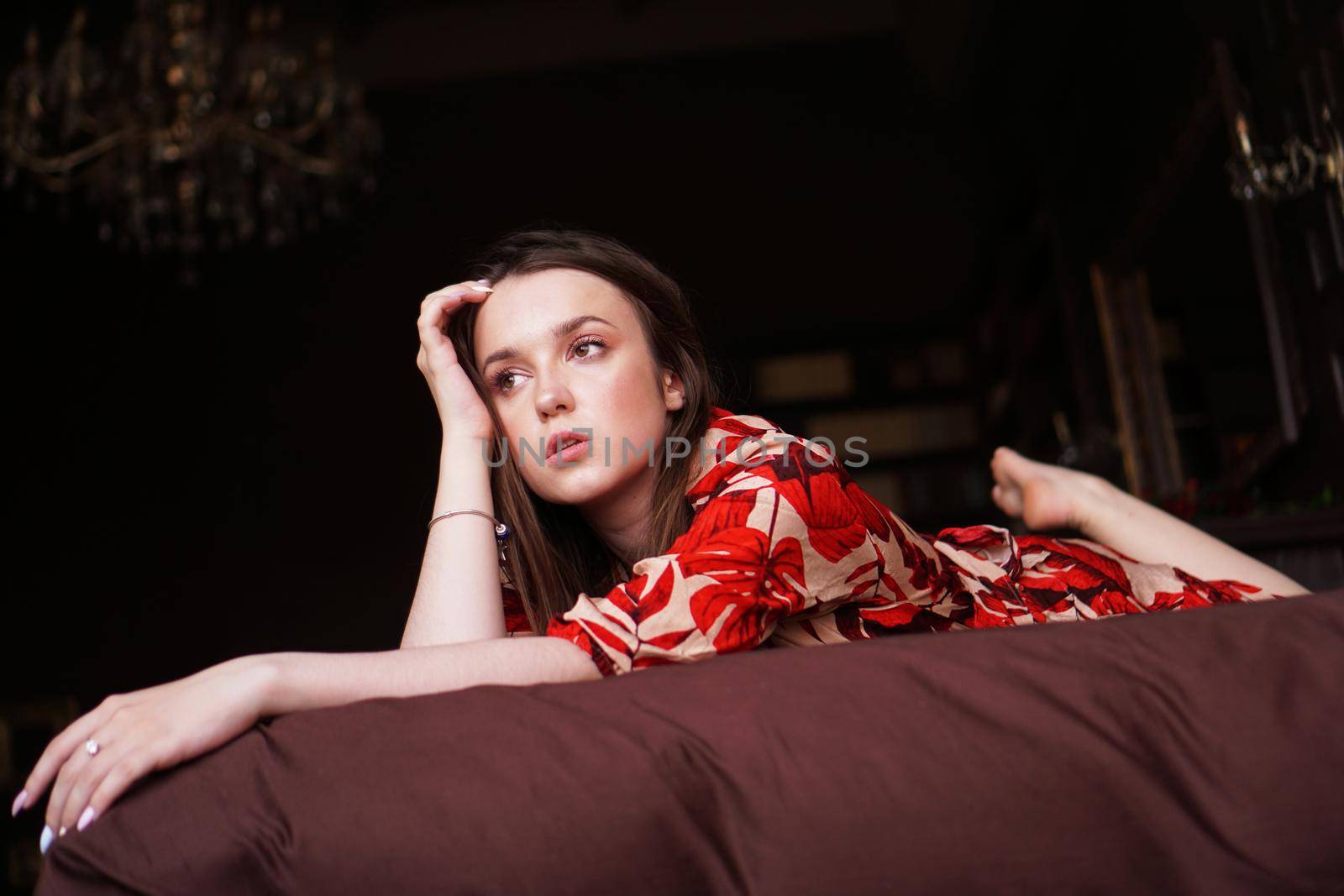 Relaxed woman lying on a brown bed thinking deeply. Woman in retro style room in red dress