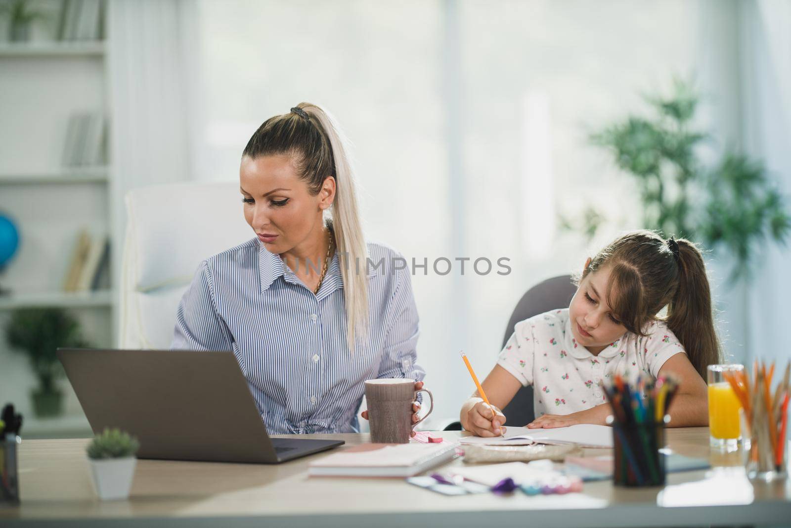 Busy single mother using laptop while her cute daughter learning at home.