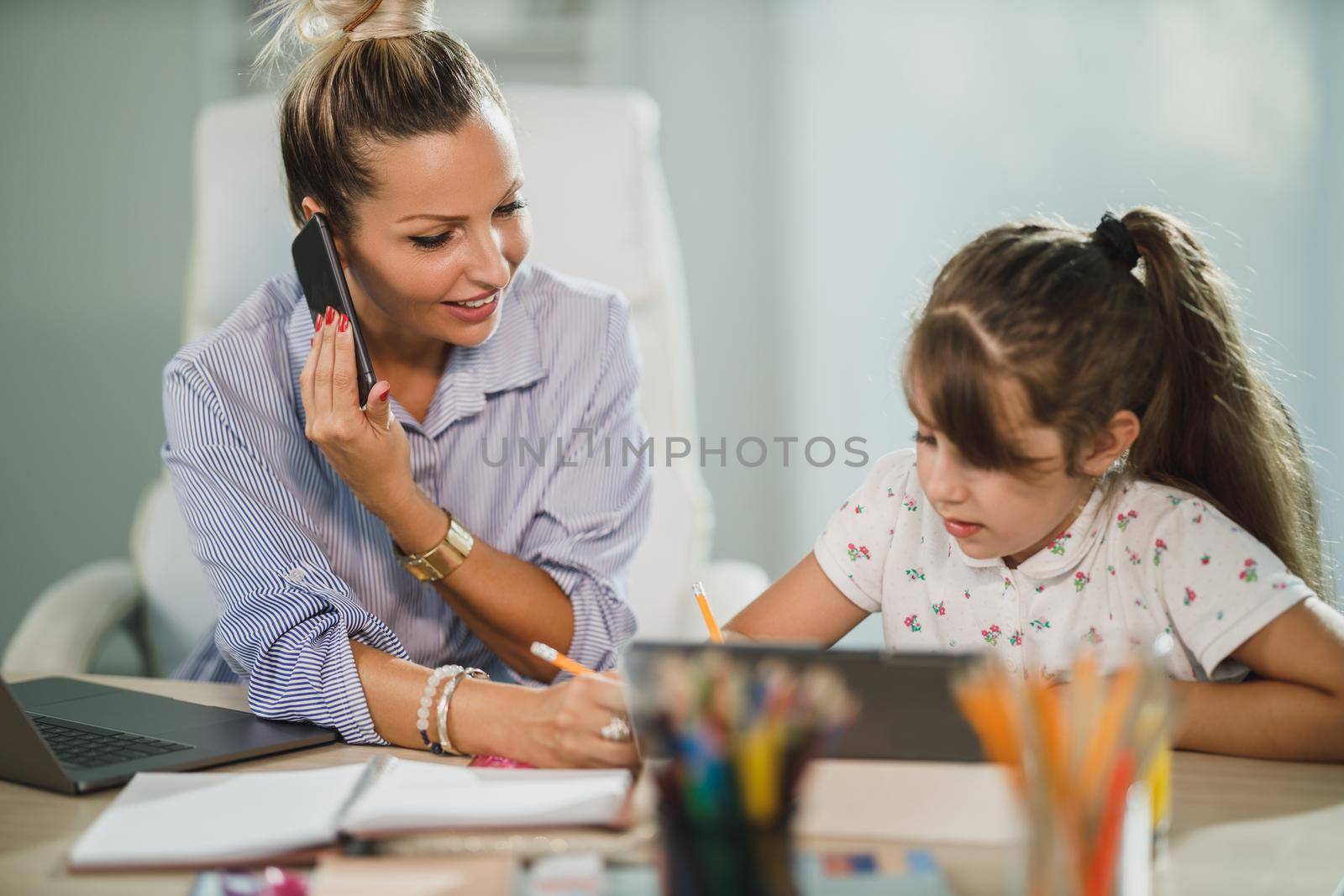 A beautiful busy single mother helping her cute daughter with homework while talking on her smartphone at home.