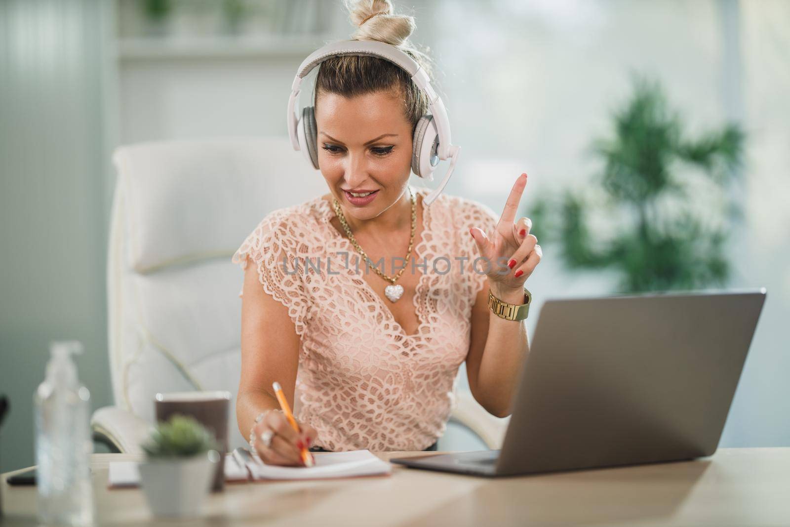 An attractive business woman with headphones sitting alone in her home office and working with client online at her laptop.