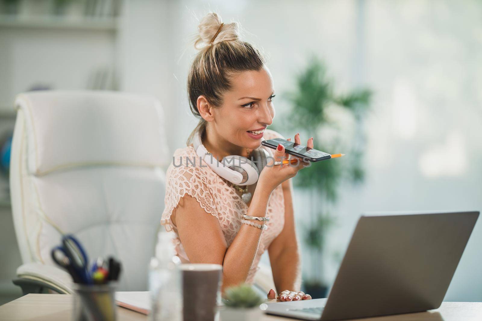 An attractive business woman sitting in her office and sending voice message on her cellphone while working online at her laptop.
