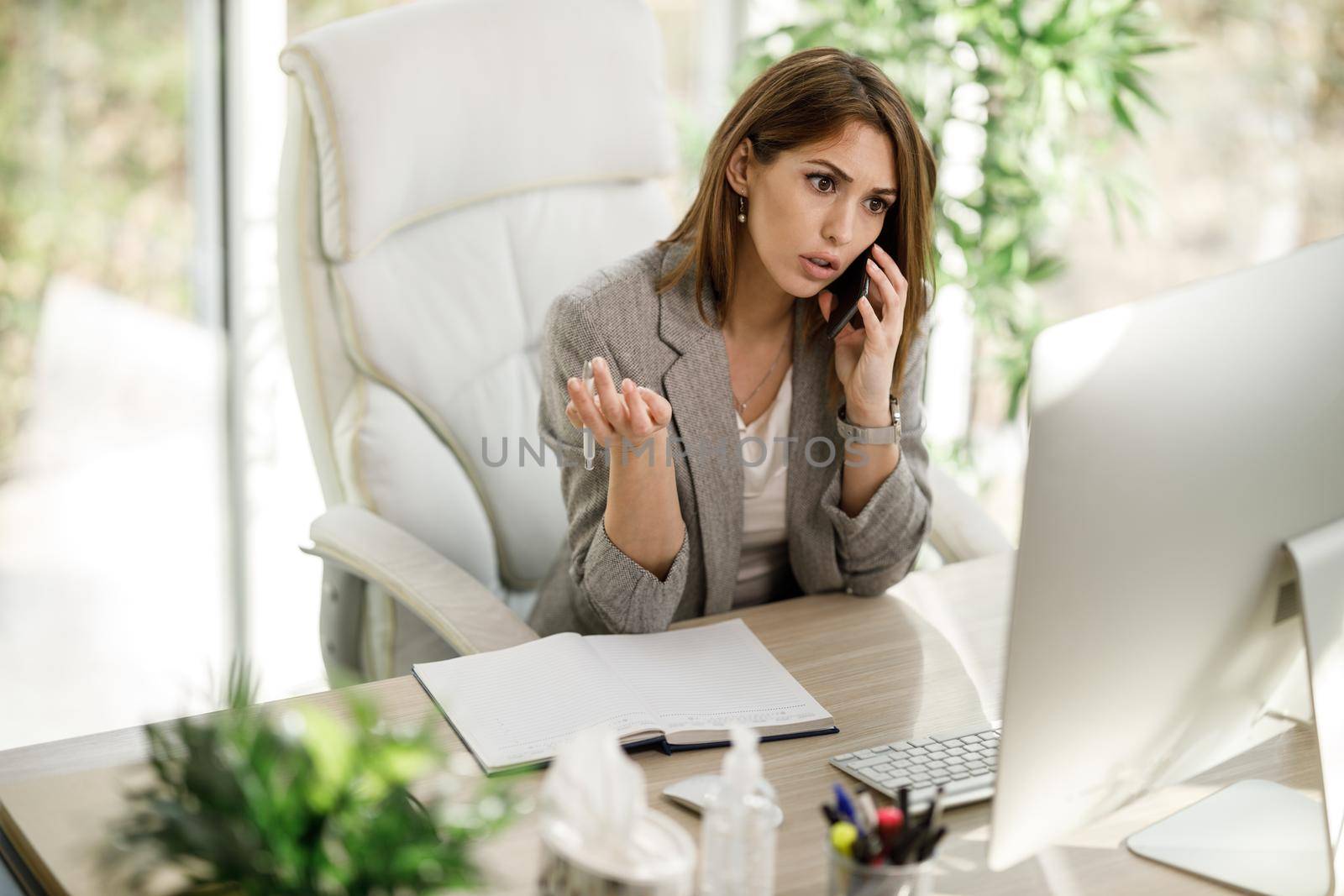 An attractive business woman sitting alone and talking on smartphone while working at desk in her home office.