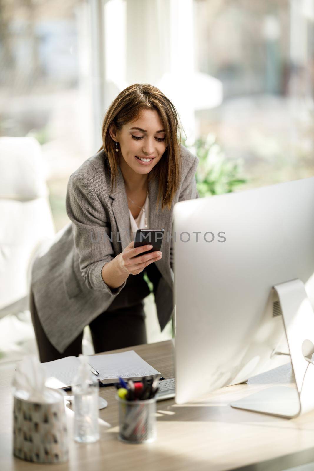 An attractive business woman using smartphone while working at computer in her office.