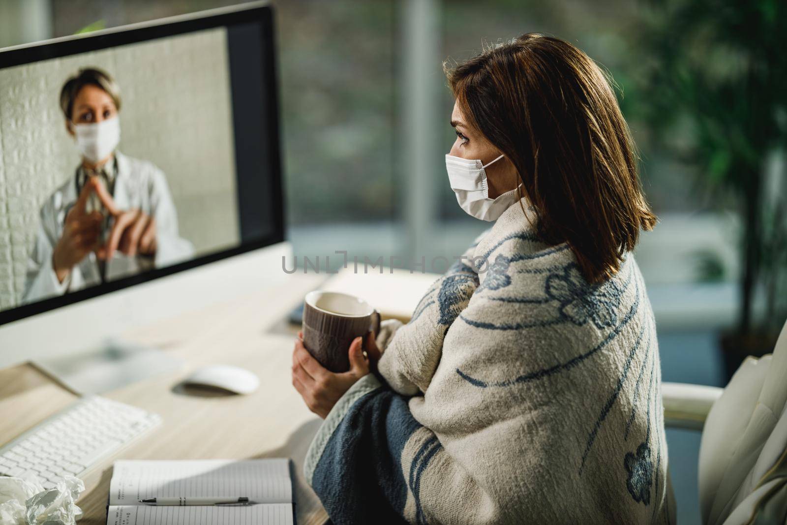 Shot of a young woman wrapped in blanket having a video call with a her doctor on a computer.