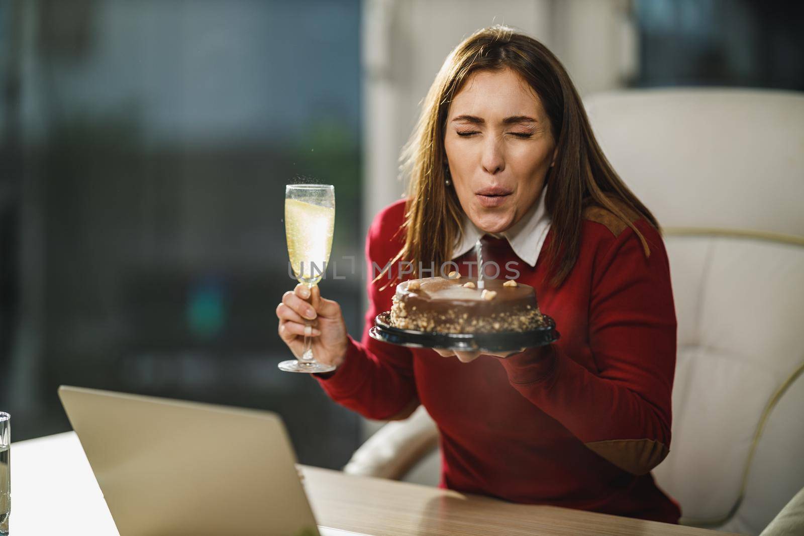 Alone young woman blowing candles on a birthday cake while have a online birthday party with friends.