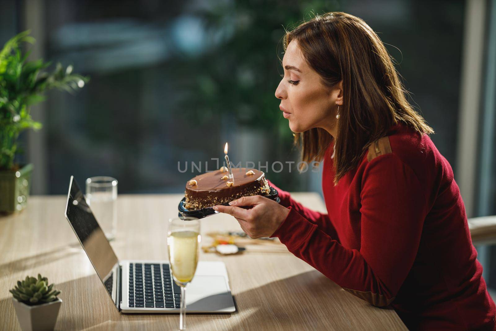 Alone young woman blowing candles on a birthday cake while have a online birthday party with friends.