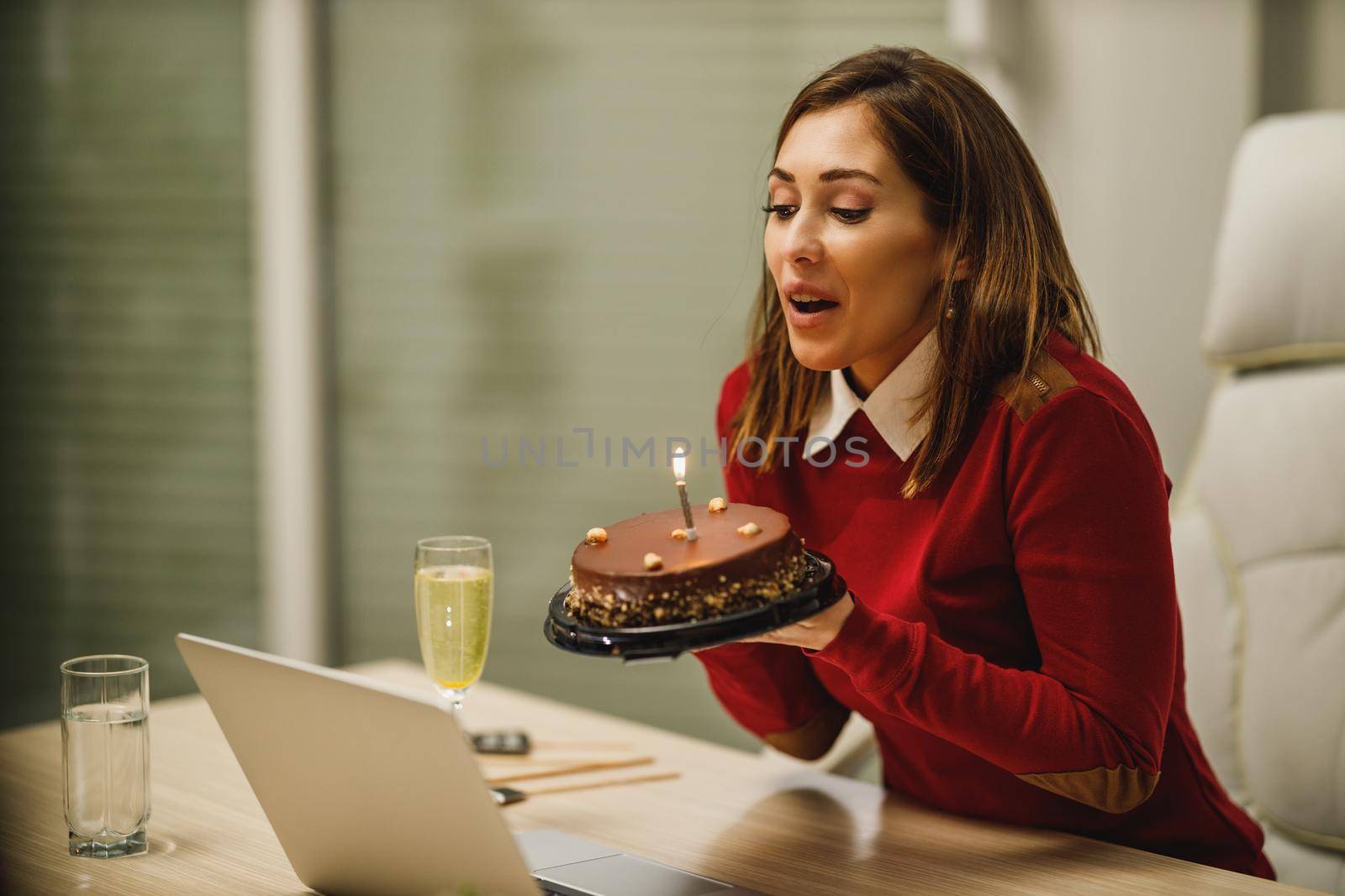 Happy young woman blowing candles on a birthday cake while have a online birthday party with friends.