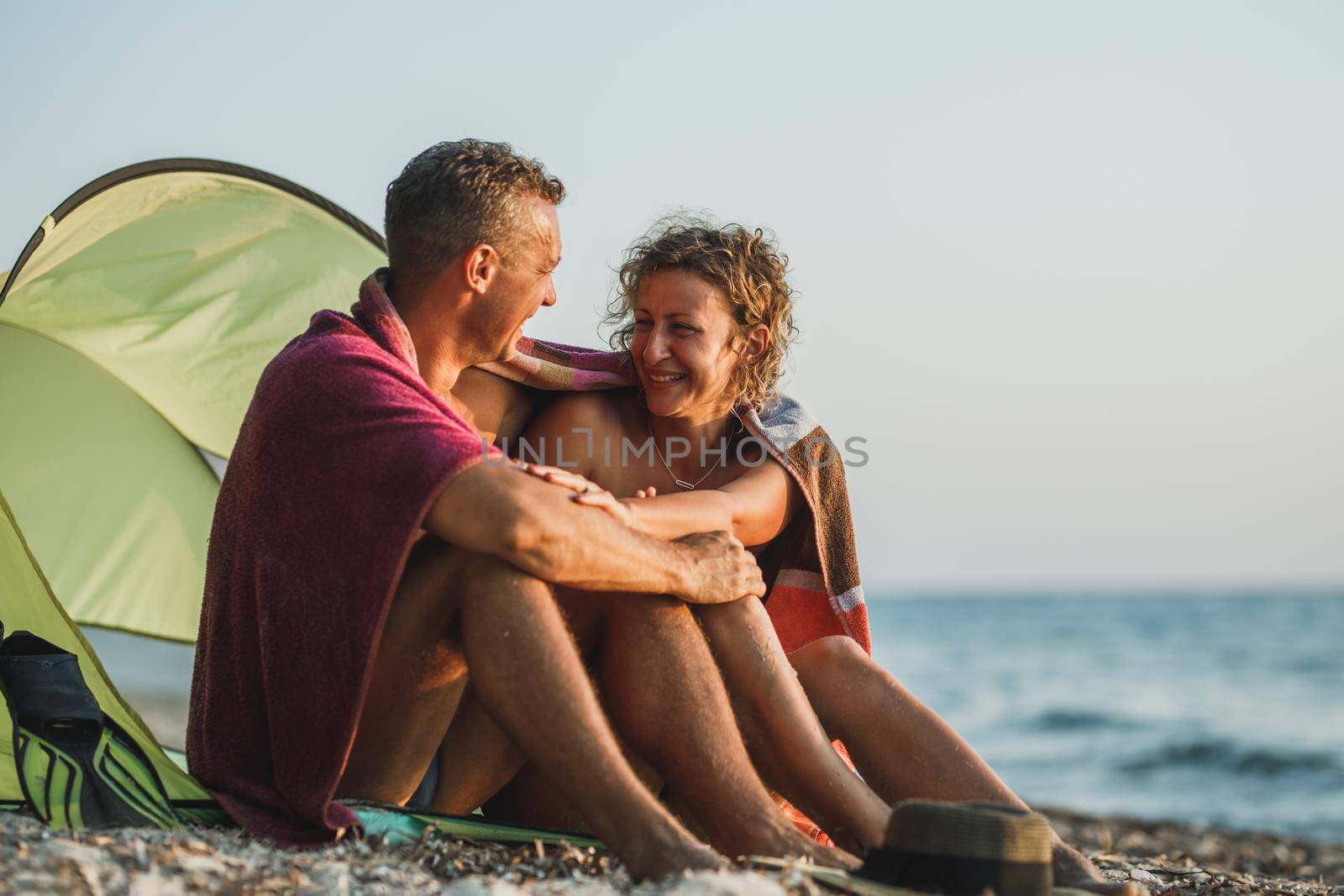 An affectionate young couple having fun at the beach and enjoying sunset wrapped in a blanket.