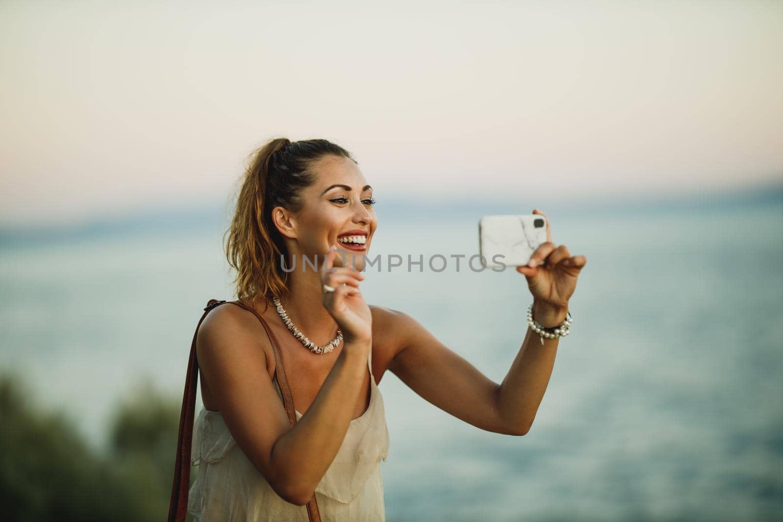 A young cheerful woman making video call on a smartphone while enjoying a summer vacation and exploring the wonderful seaside of Meditteranian.