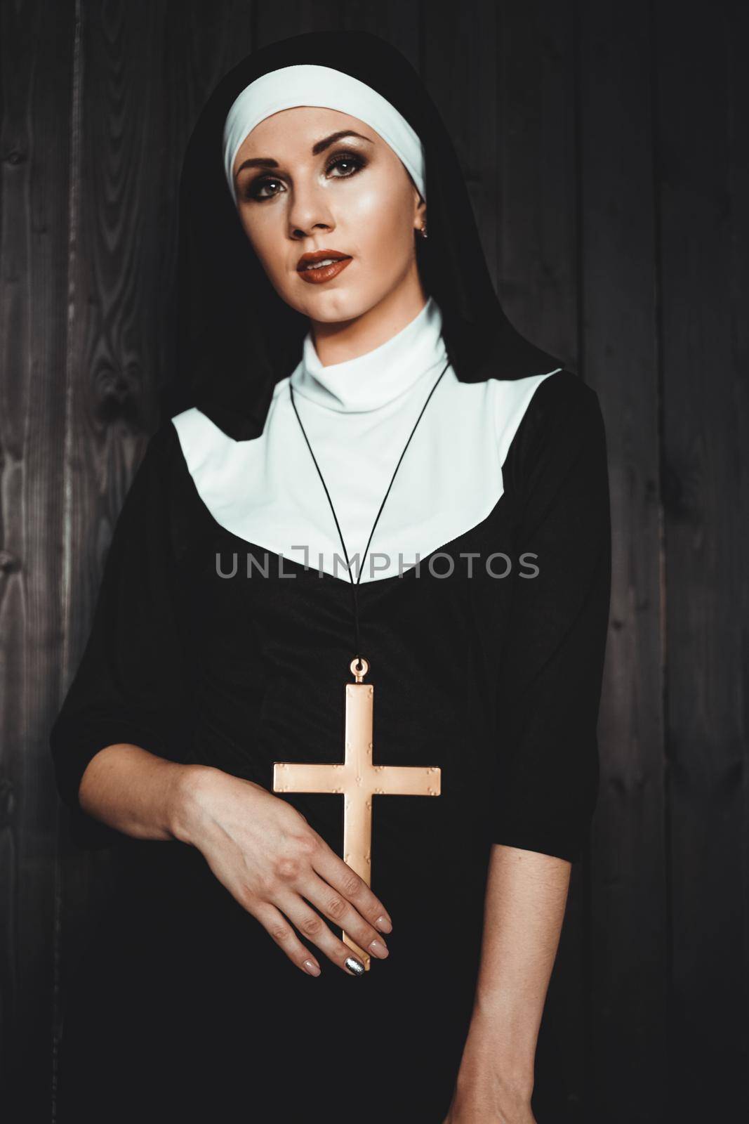 Sexy nun prays indoor . Beautiful young holy sister. Young beautiful nun with a cross in a robe in a black interior.