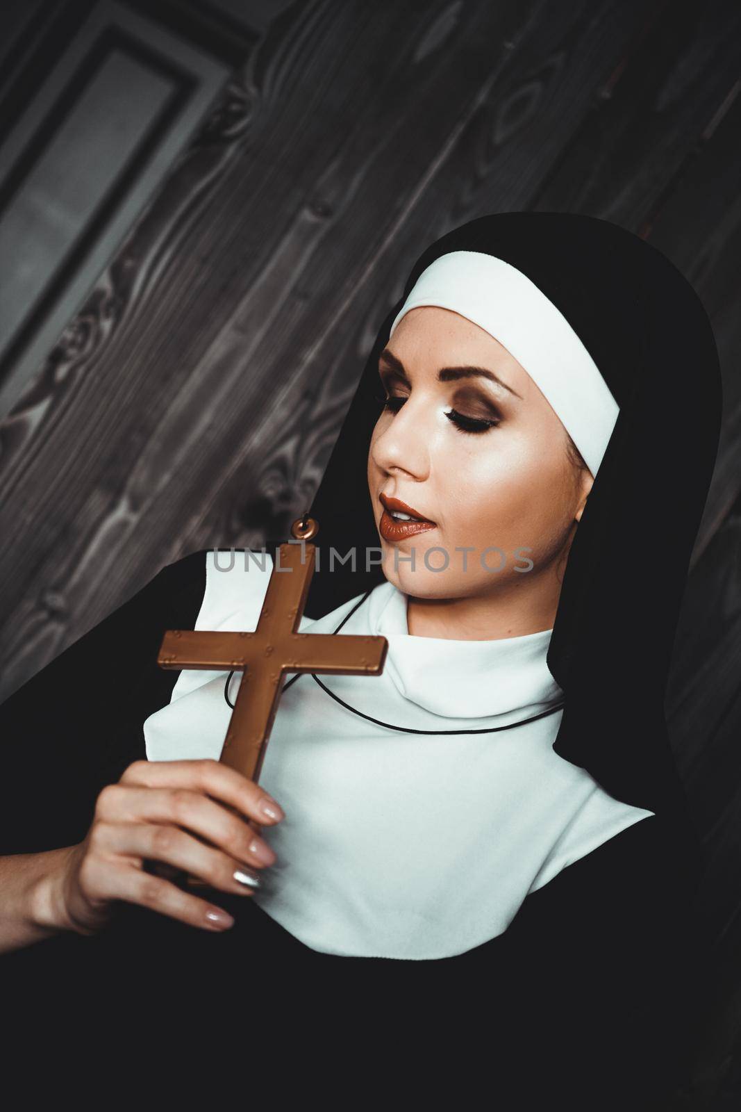 Sexy nun prays indoor . Beautiful young holy sister. Young beautiful nun with a cross in a robe in a black interior.