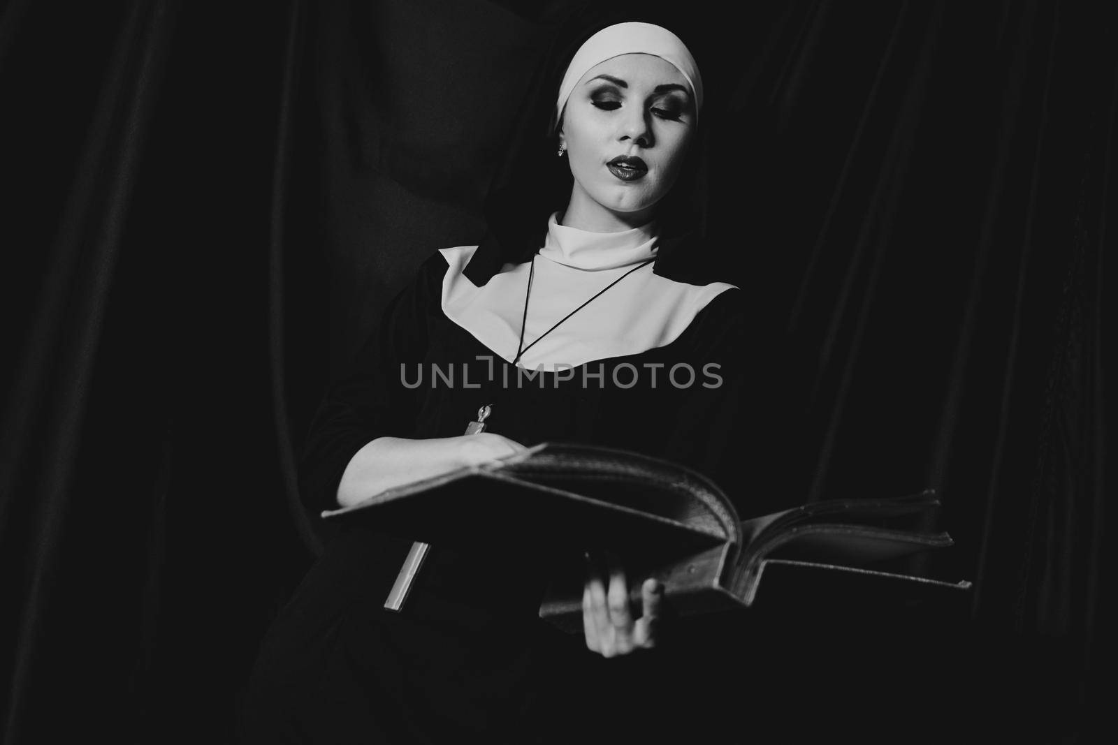 Beautiful young nun in religion black suit holds Bible and posing on camera with big book on a black background. Religion concept. Black and white