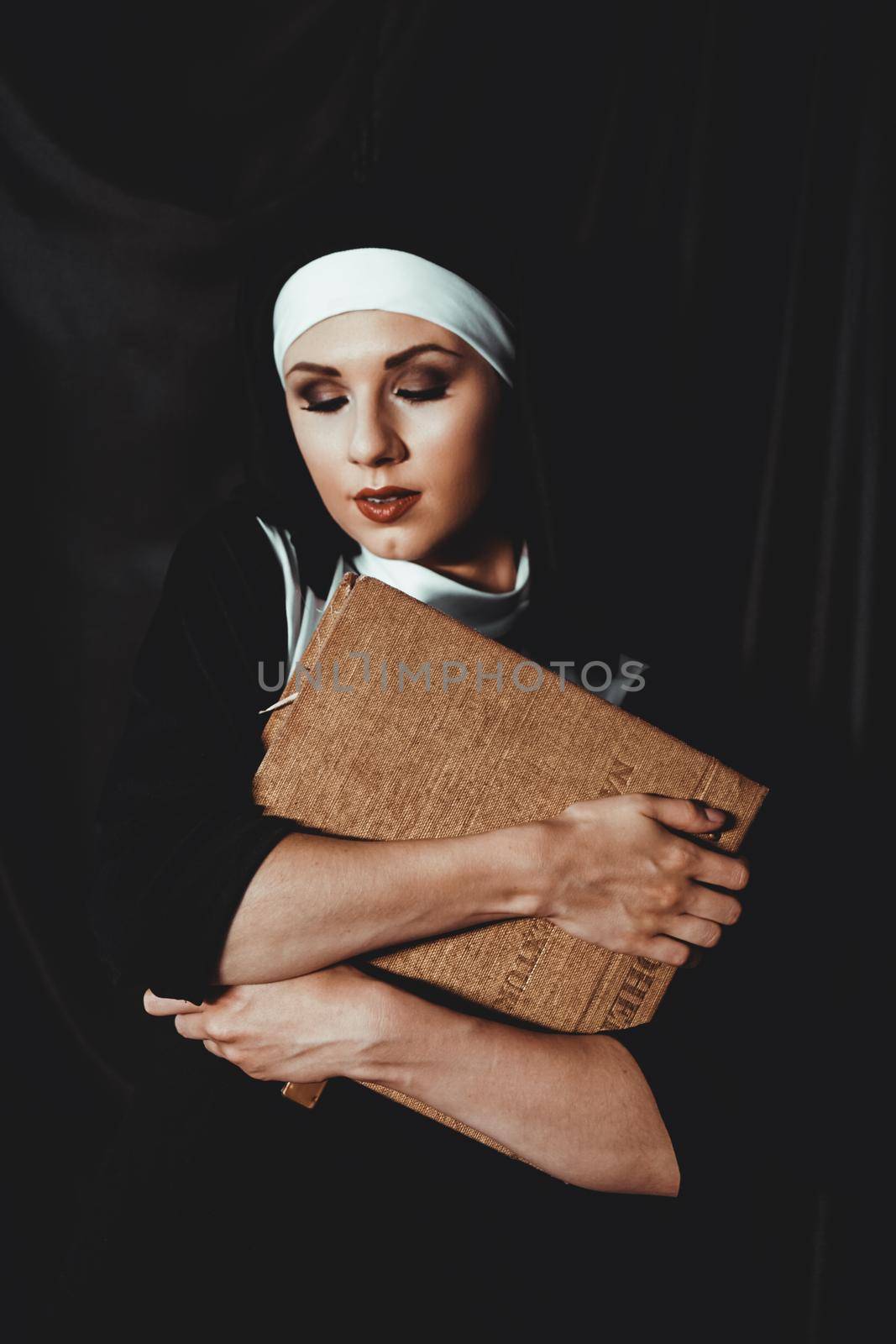 Beautiful young nun in religion black suit holds Bible and posing on camera with big book on a black background. Religion concept.