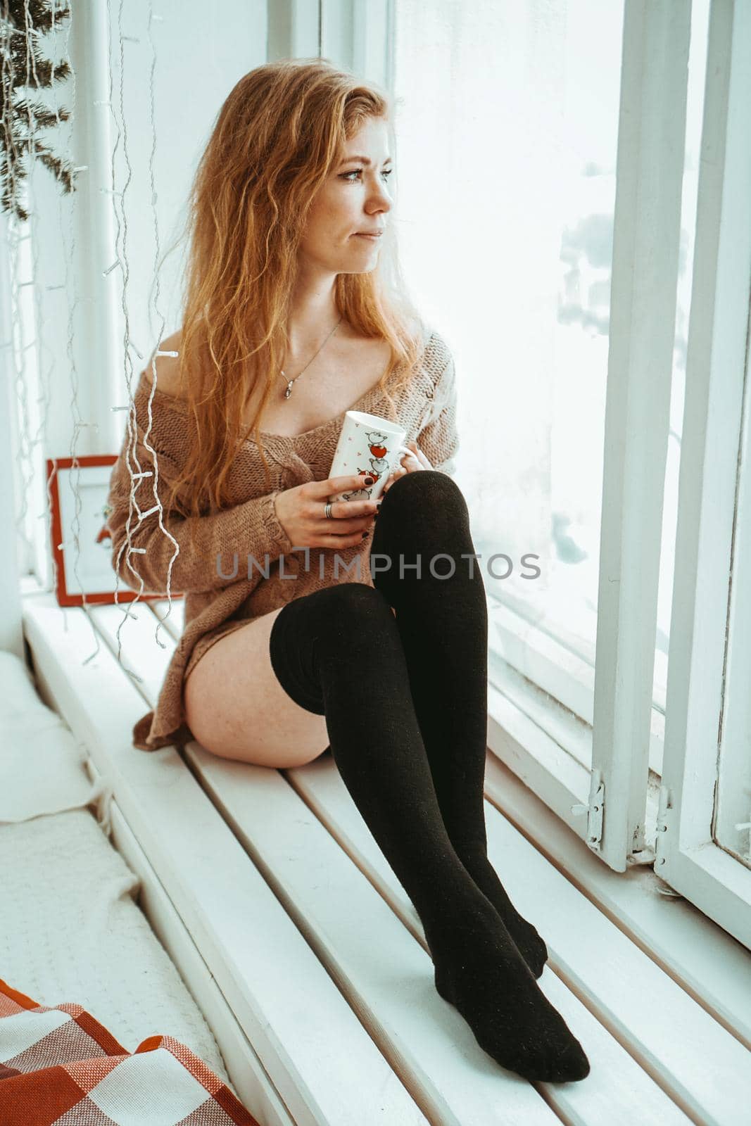 Beautiful young woman drinking coffee and looking through window while sitting at windowsill at home