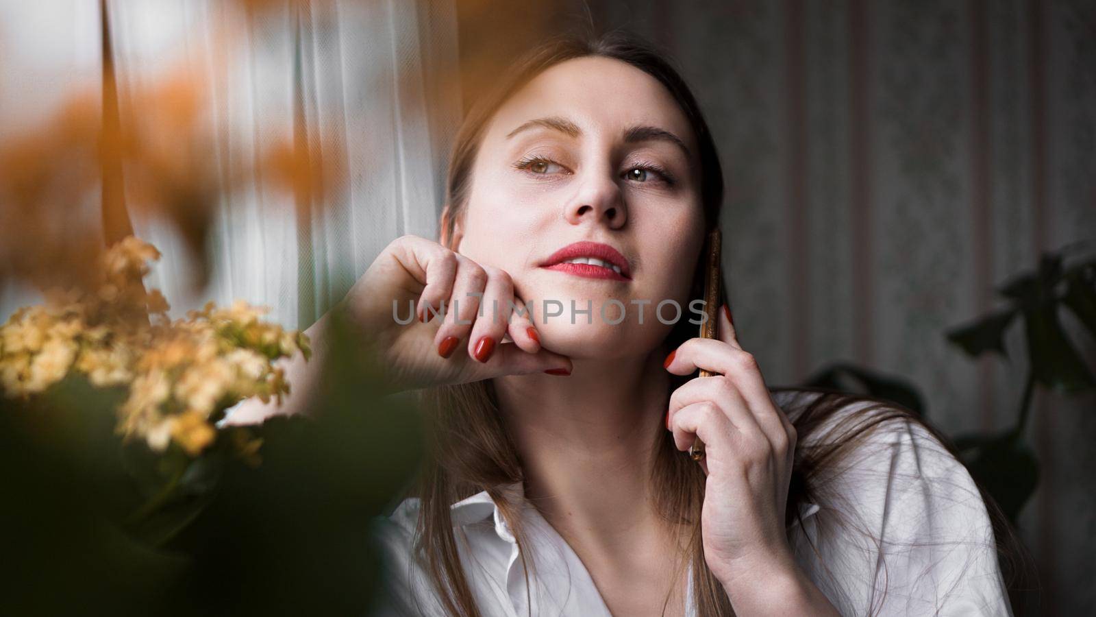 Pretty woman with red lips talking on phone near window at home in morning sunlight