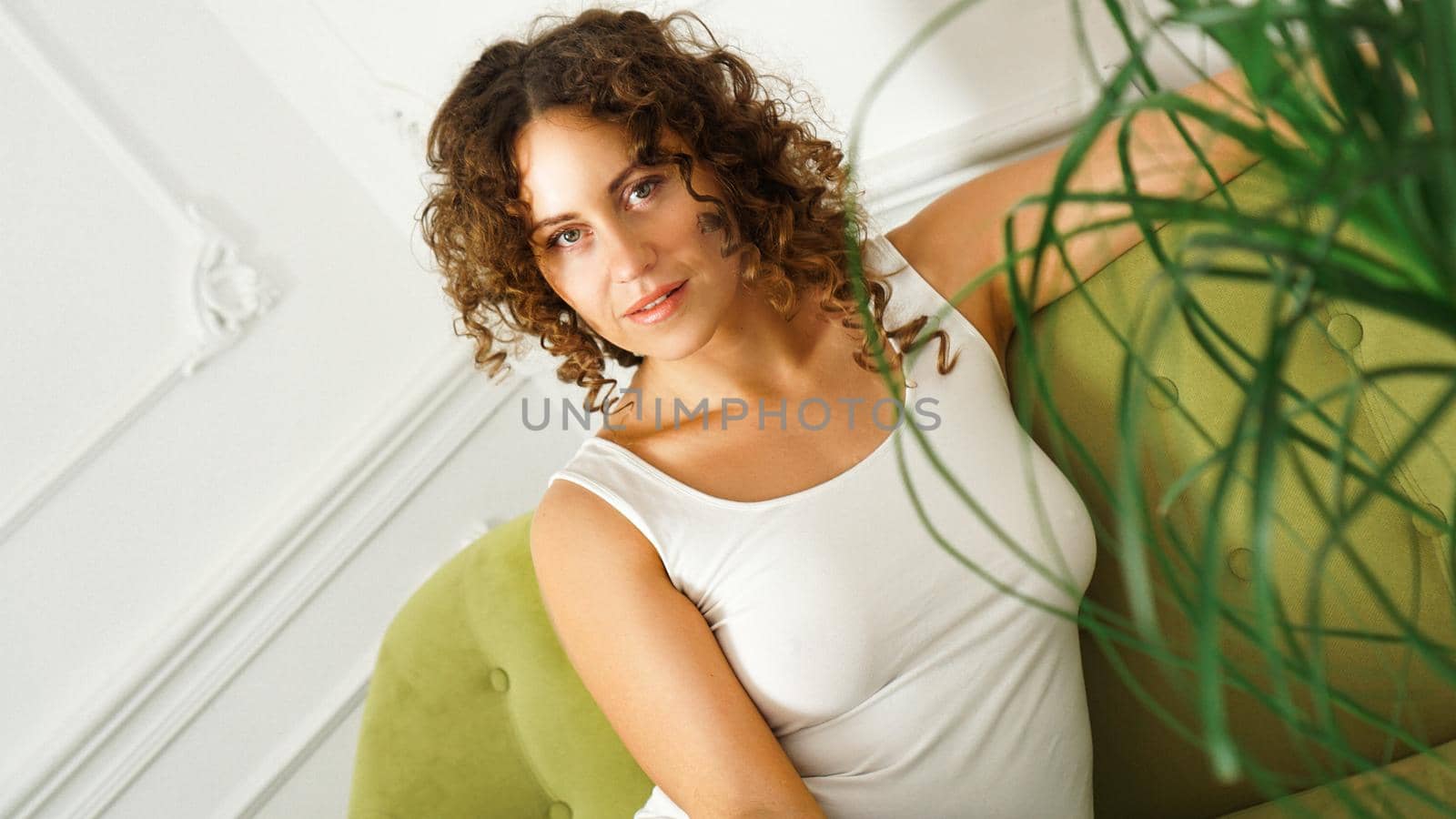 Relaxed lifestyle. Happy young woman with curly hair in white t-shirt spending time at home. Green sofa and white walls in room