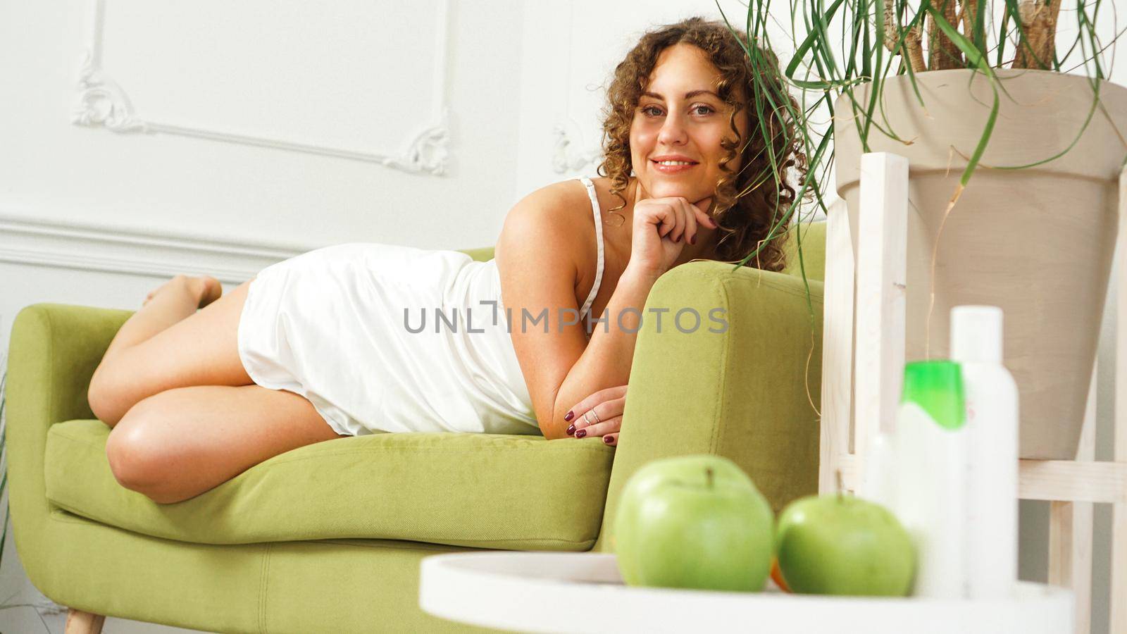 Beauty cosmetics concept. Beautiful young woman with curly hair and healthy skin in room with a green sofa. Apples and Organic Care on a wooden table