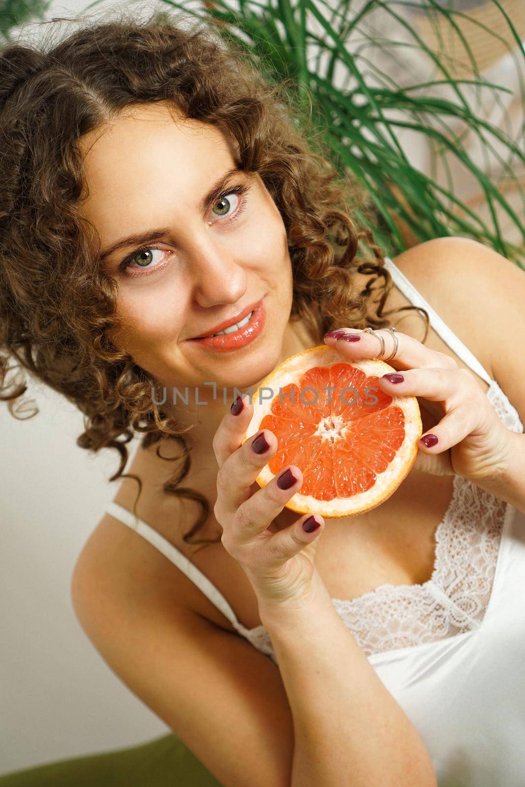 Portrait of pretty middle-aged woman with curly hair with grapefruit at home - light room. Happiness, beauty and health - vertical photo