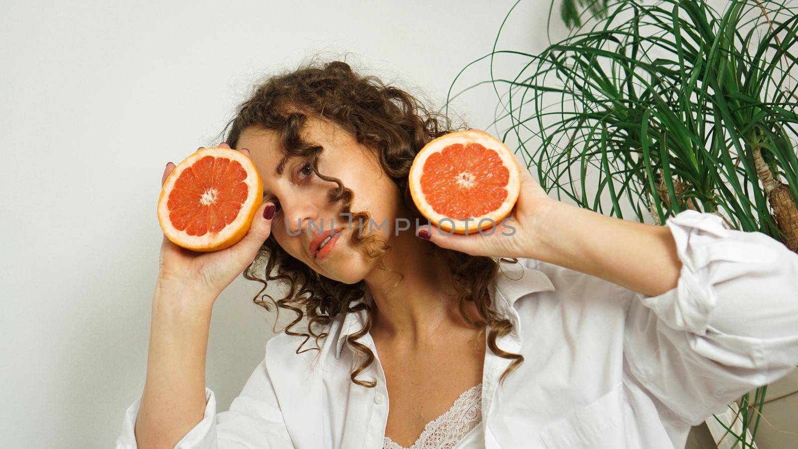 Portrait of pretty middle-aged woman with curly hair with grapefruit at home - light room. The concept of art, beauty and health